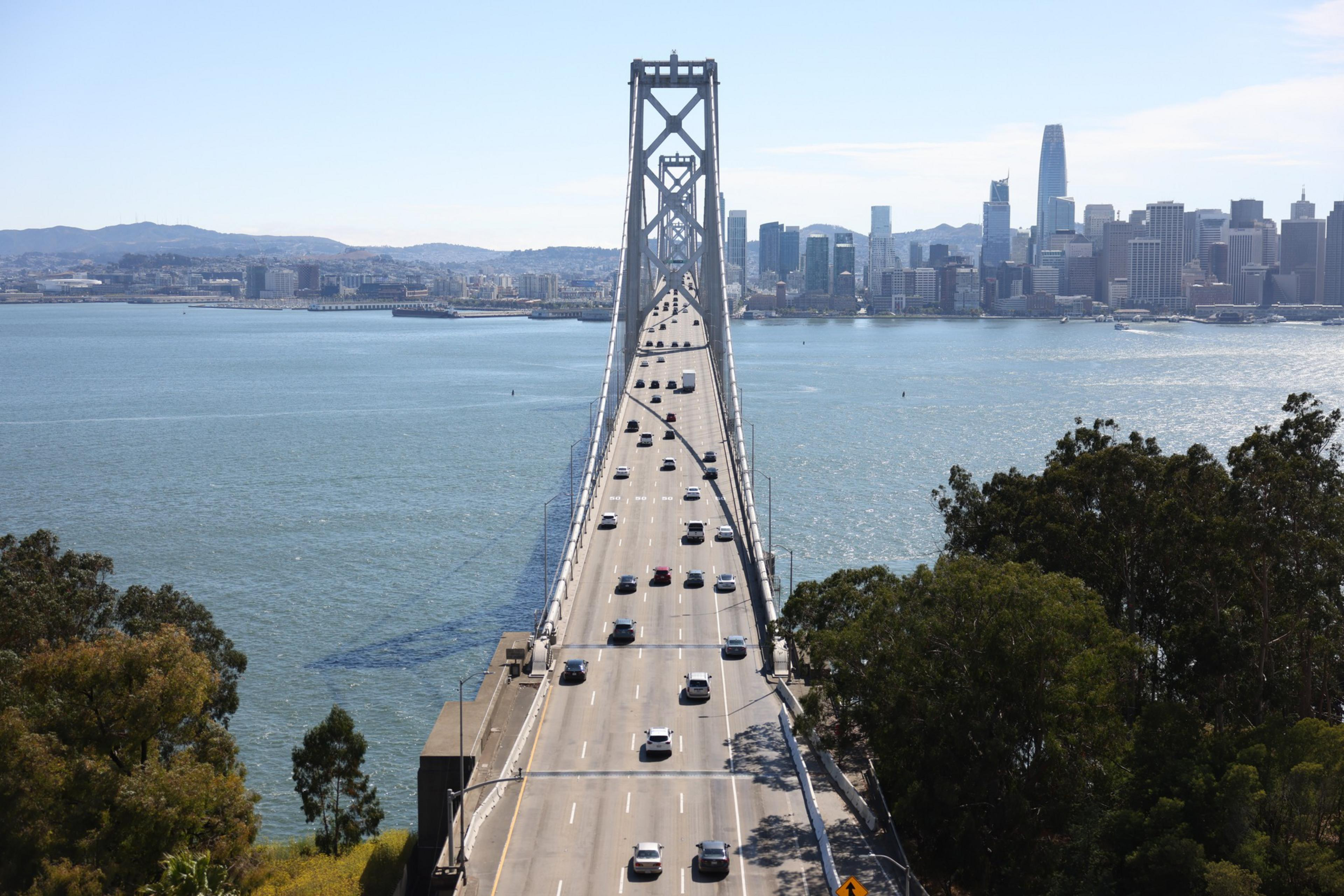 A large suspension bridge stretches over water with cars traveling in both directions, connecting to a city skyline with tall buildings in the background.