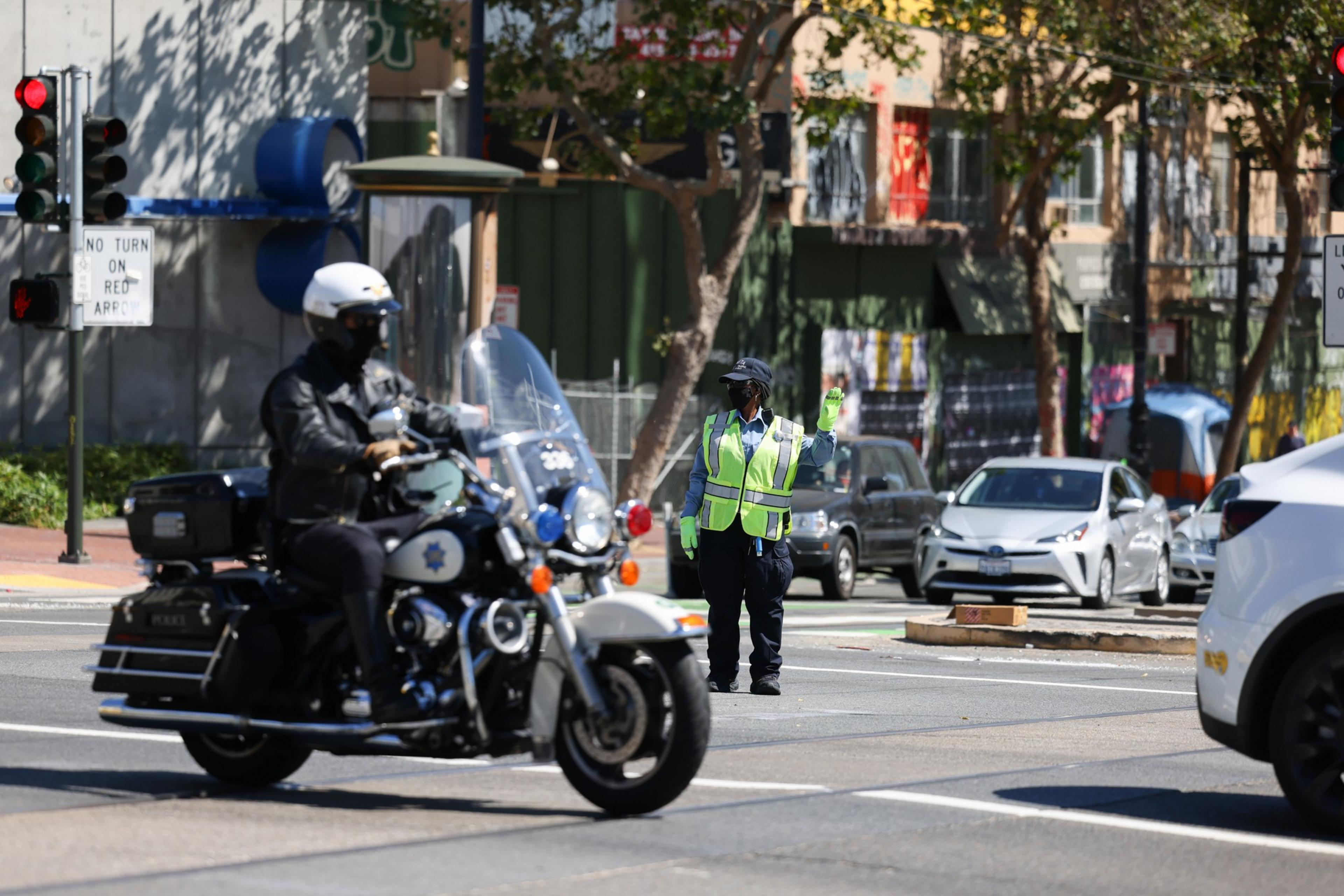 A police officer on a motorcycle rides past a traffic controller in a neon vest directing vehicles at a city intersection with traffic lights and a no turn sign.