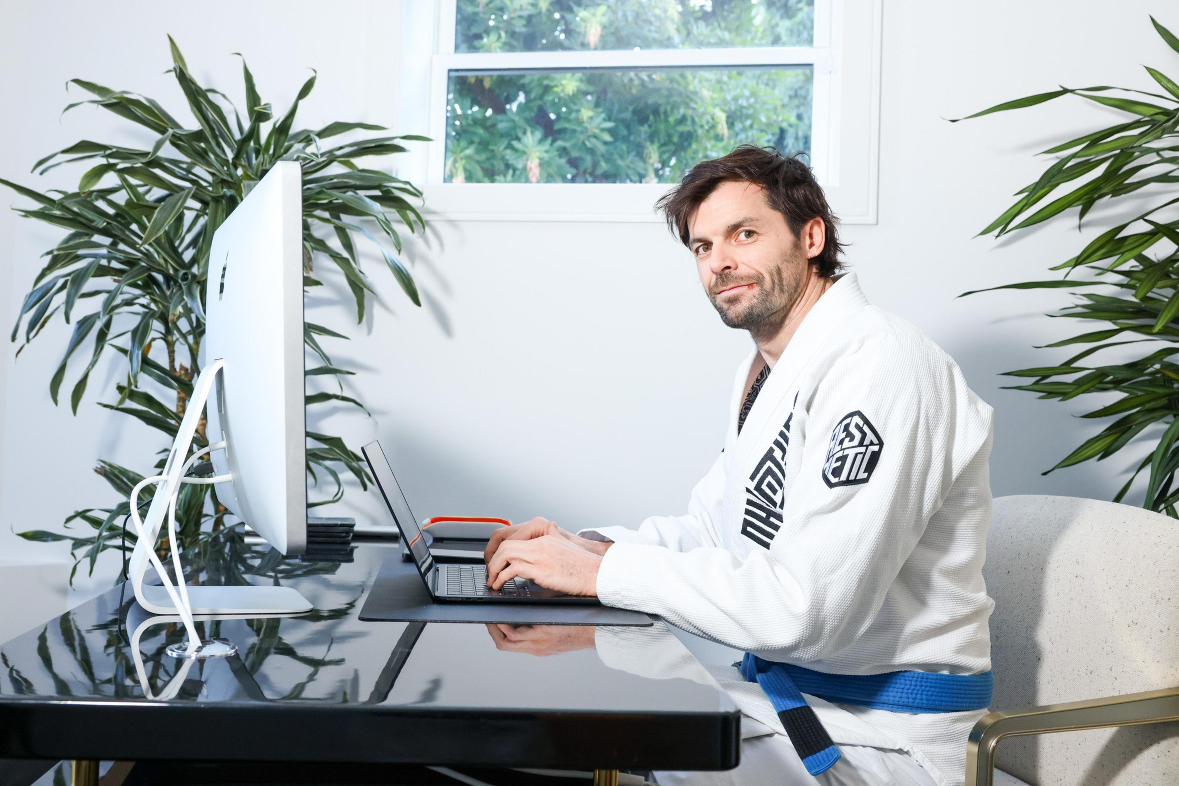 A person in a white martial arts gi with a blue belt sits at a sleek black desk, working on a laptop. Two large plants flank the desk, and a window shows greenery outside.