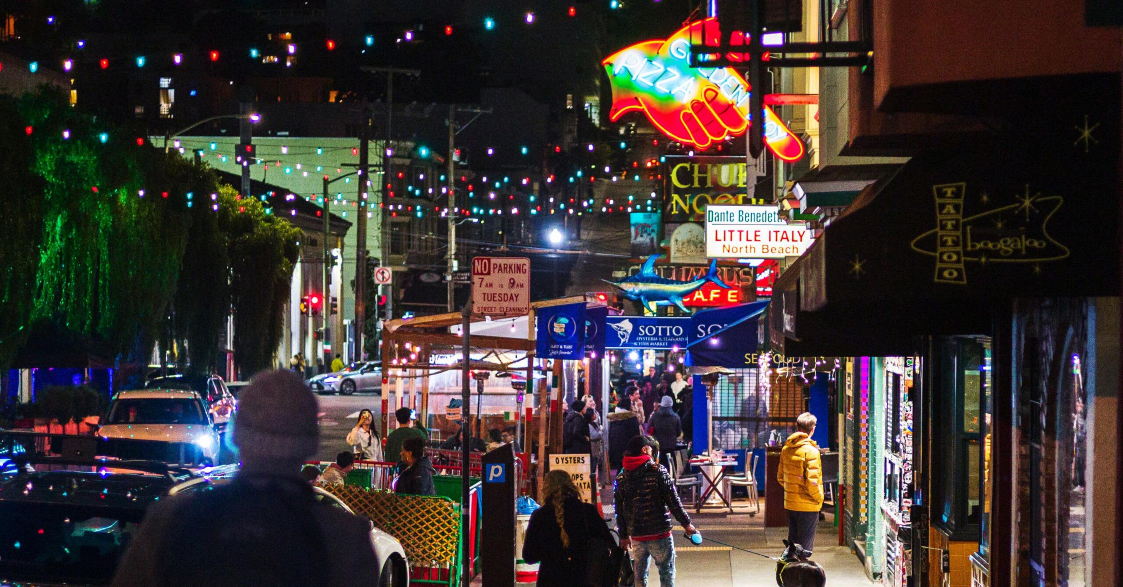 A vibrant evening street scene shows colorful string lights, bustling people, neon signs for pizza and cafes, and a lively atmosphere with cars and pedestrians.