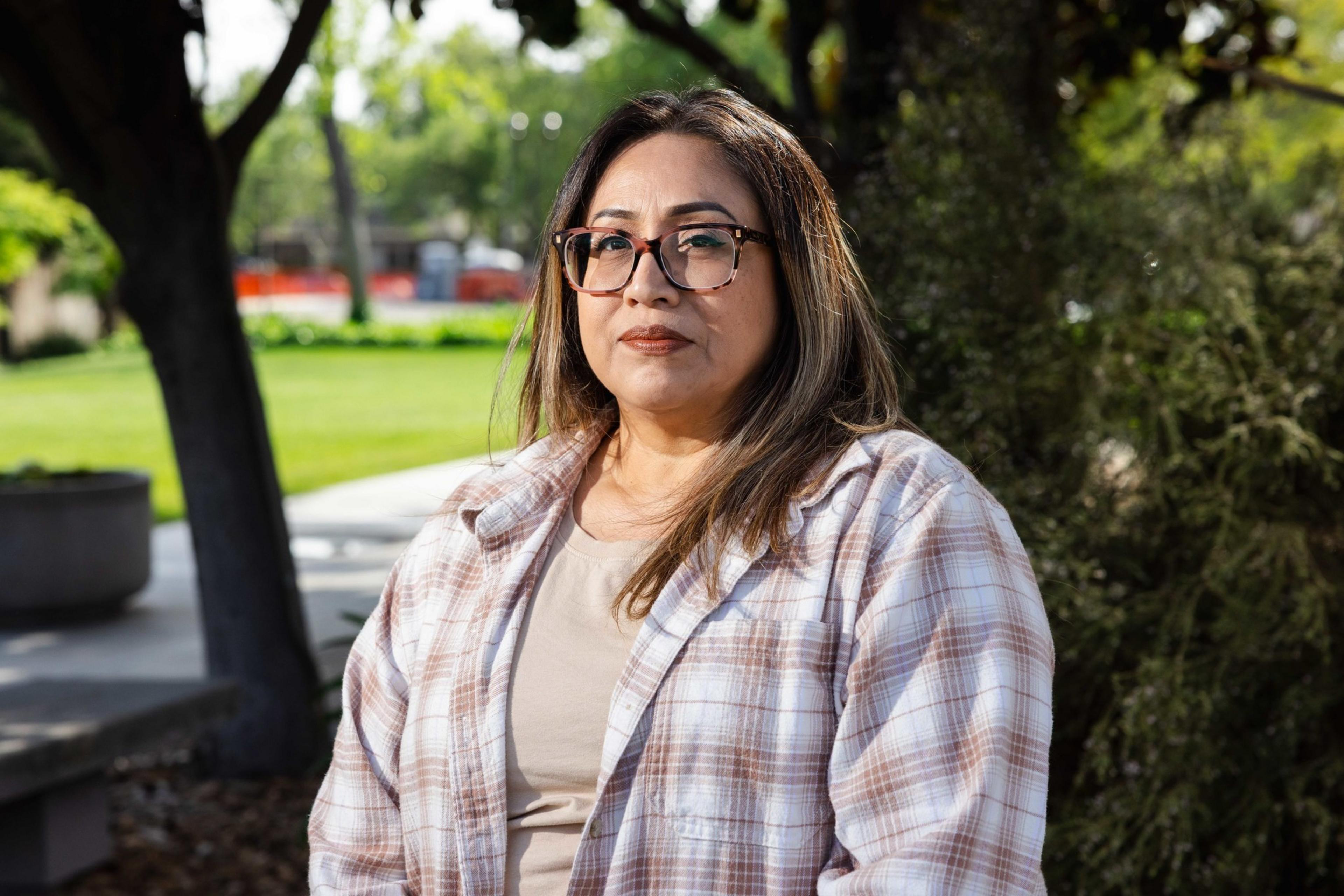 A woman with glasses and long hair stands in a park, wearing a plaid shirt over a beige top. There are trees and greenery in the background.