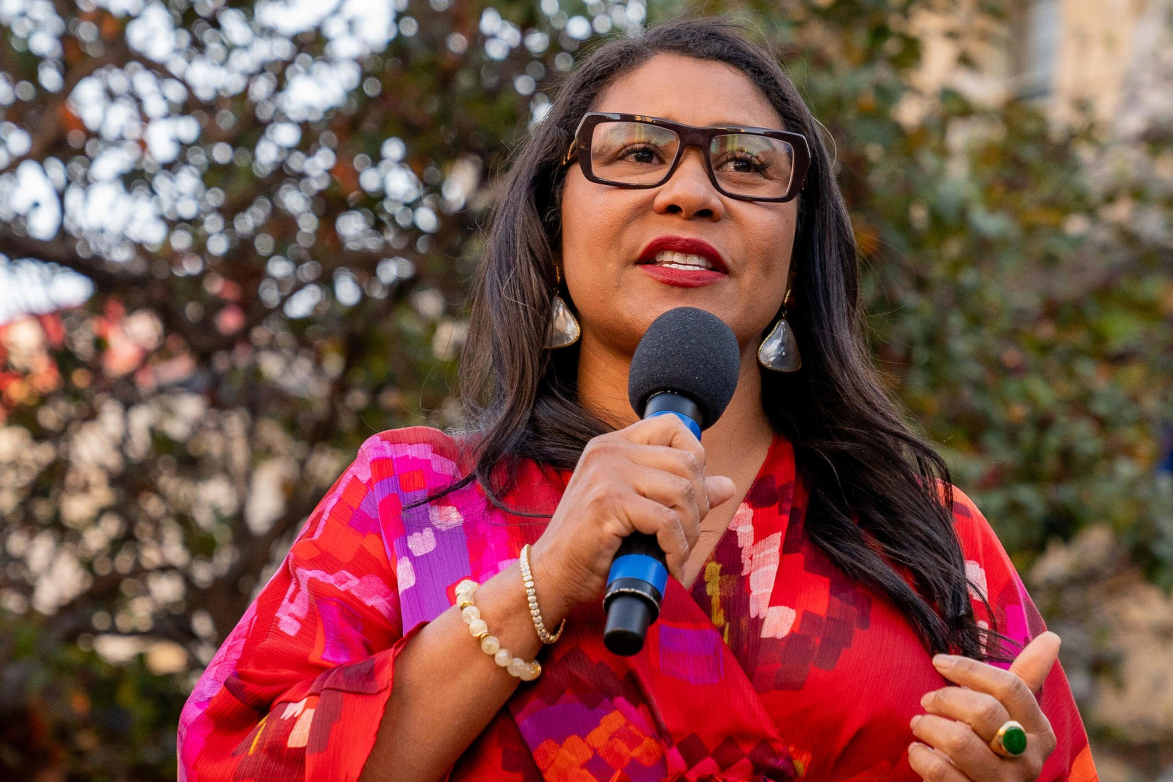 A woman with long dark hair and glasses speaks into a microphone, wearing a vibrant red and purple patterned outfit and pearl accessories.
