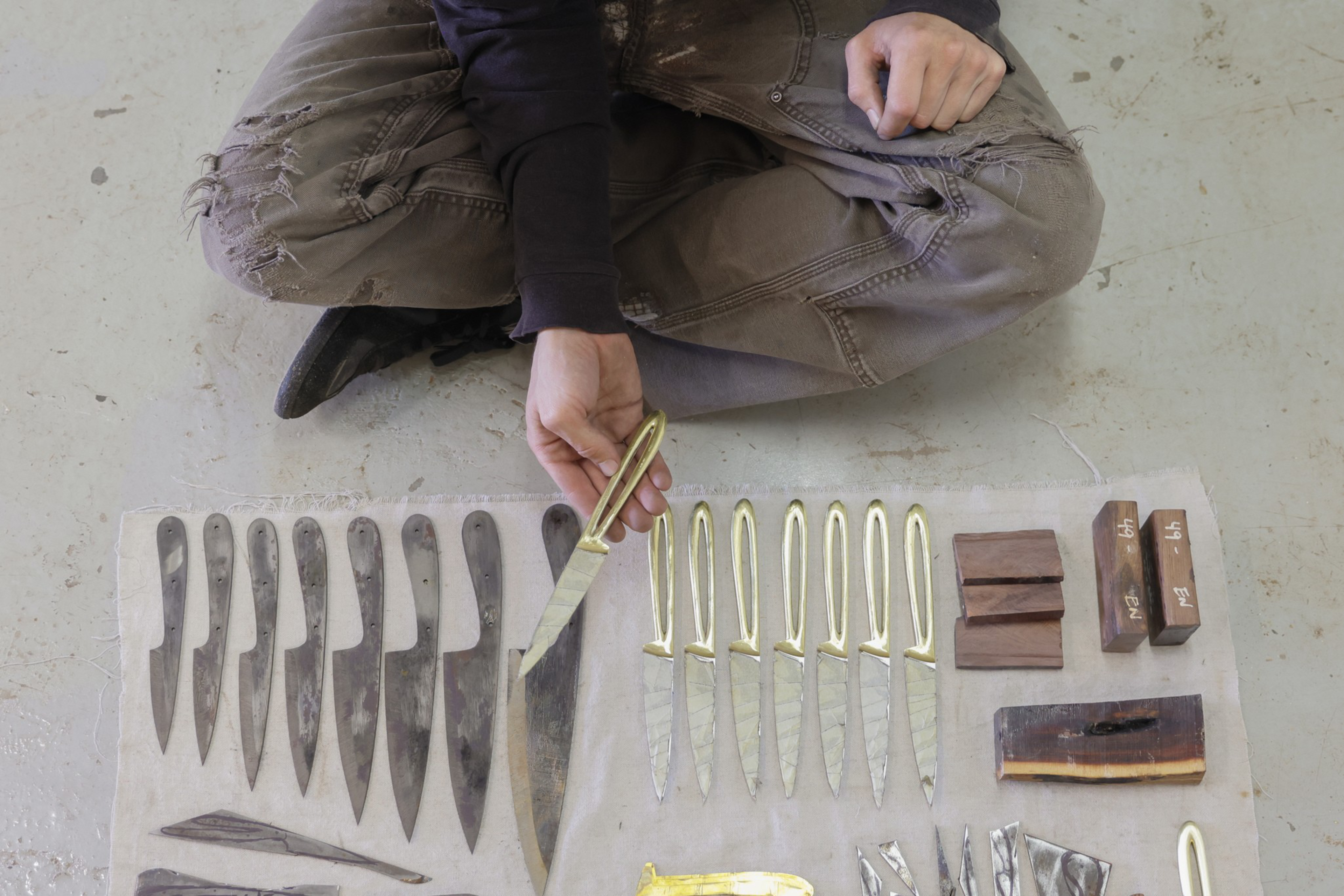 A person sits cross-legged on the floor, holding a knife with a brass handle above a cloth displaying multiple knife blades, handles, and wooden blocks.