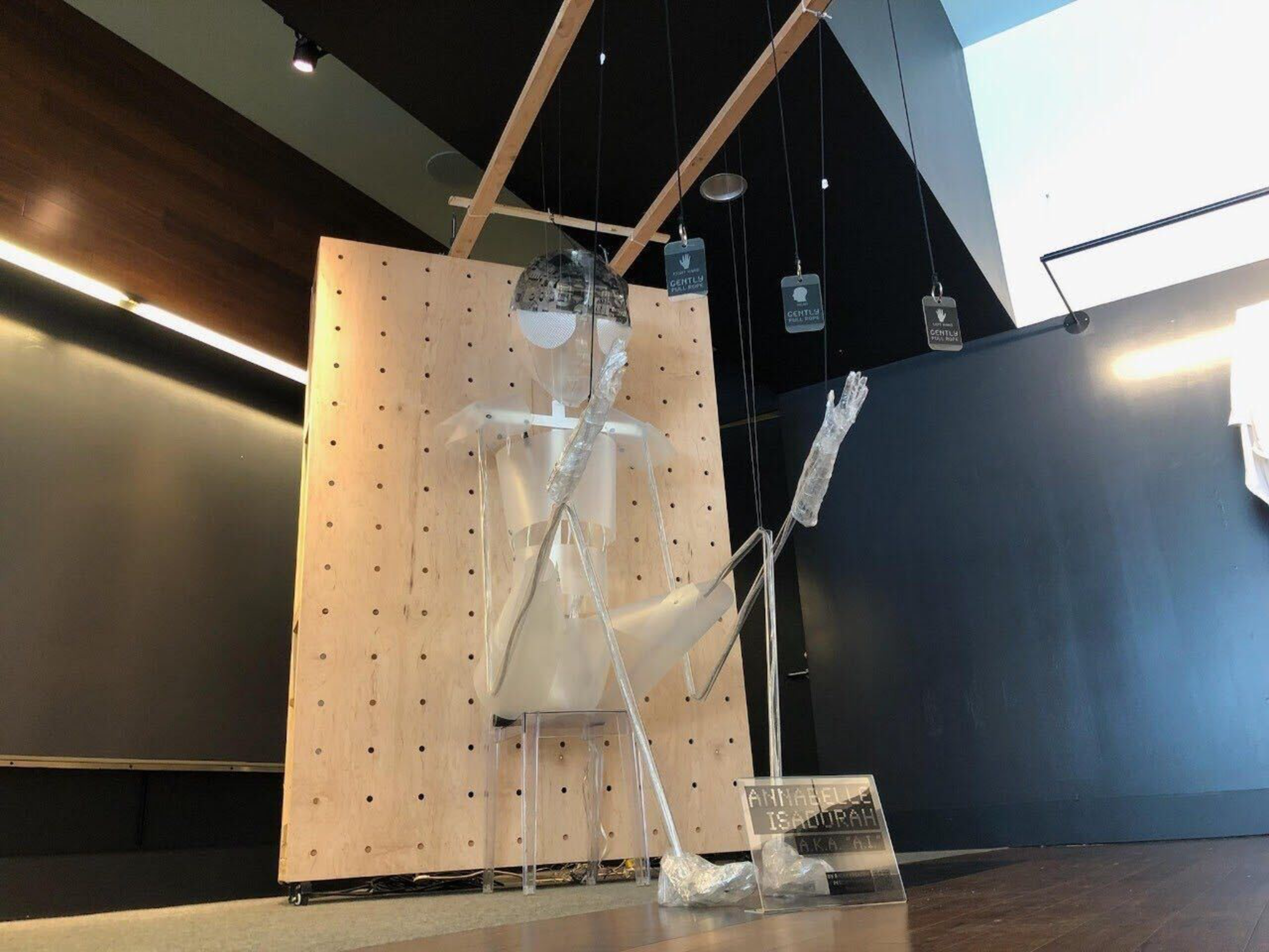 A transparent mannequin with aluminum foil-wrapped head and arms sits and waves, suspended by strings in front of a pegboard in a dark room.