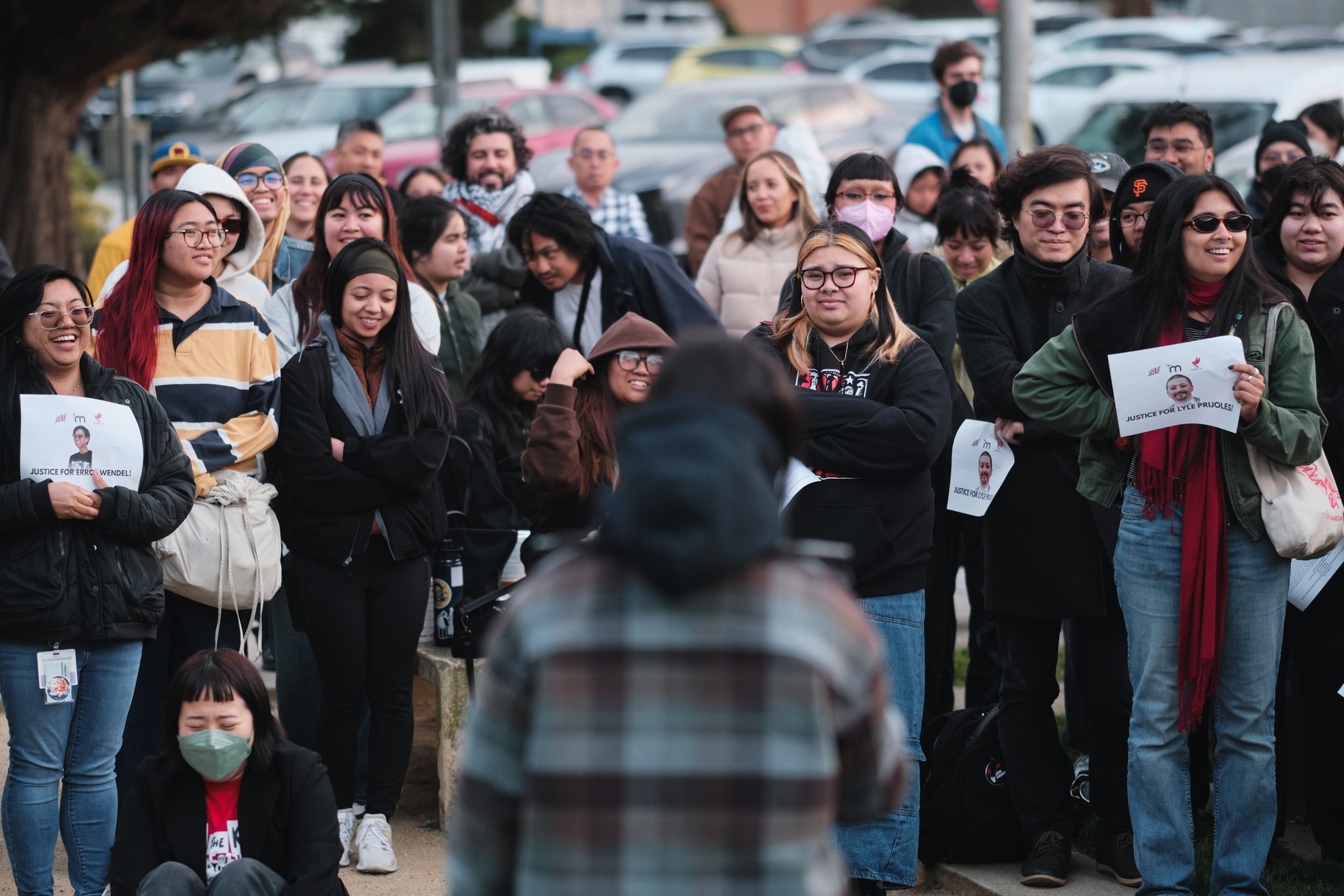 A diverse group of people stands and listens attentively, some holding signs that read “Justice for Eric Prude,” suggesting a peaceful gathering or protest.