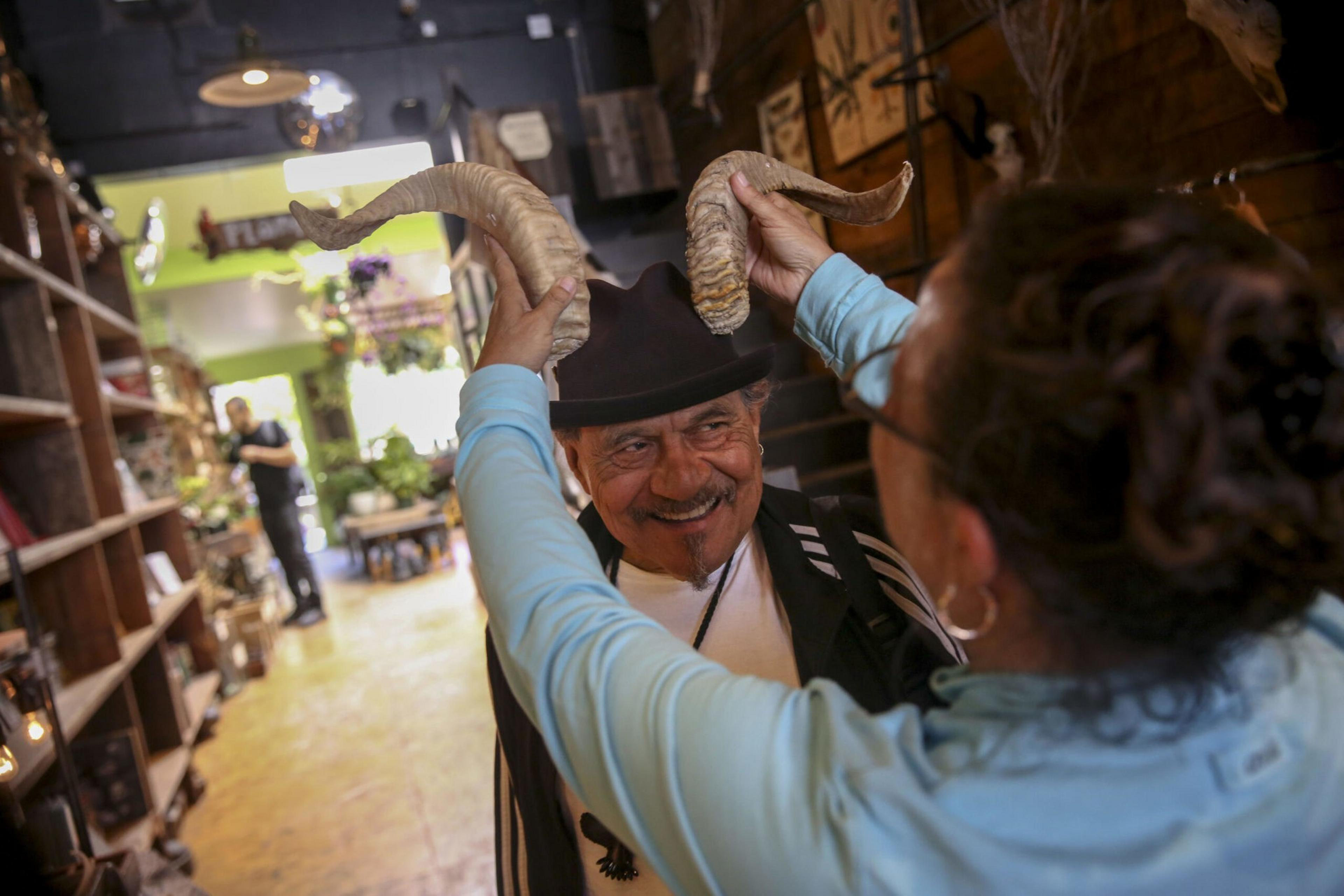 A smiling man wearing a black hat holds large curved horns above his head, while another person in a light blue jacket helps him inside a rustic shop.