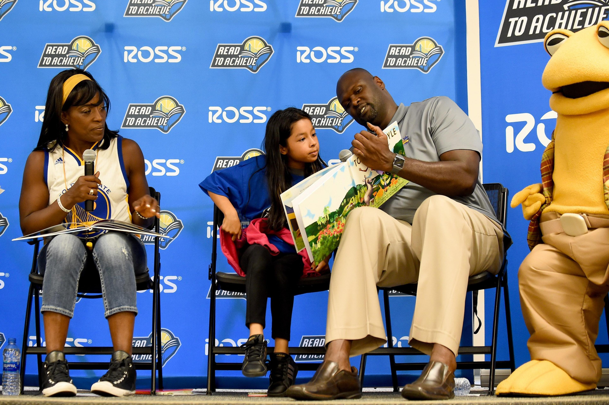 A man reads a book to a young girl sitting beside him, while a woman holds a microphone, and a mascot stands on the right, all in front of a &quot;Read to Achieve&quot; backdrop.