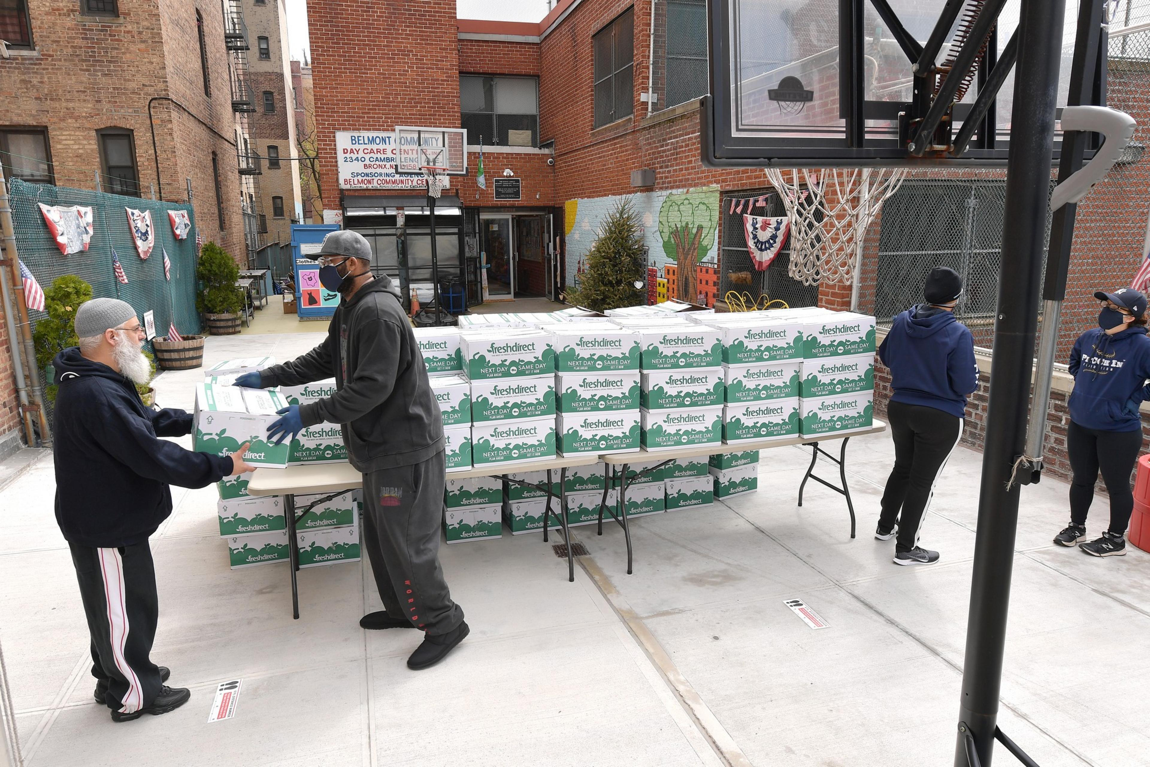 A man hands a box to another person at a table stacked with &quot;FreshDirect&quot; boxes. They're outside a brick building with a &quot;Belmont Community&quot; sign.