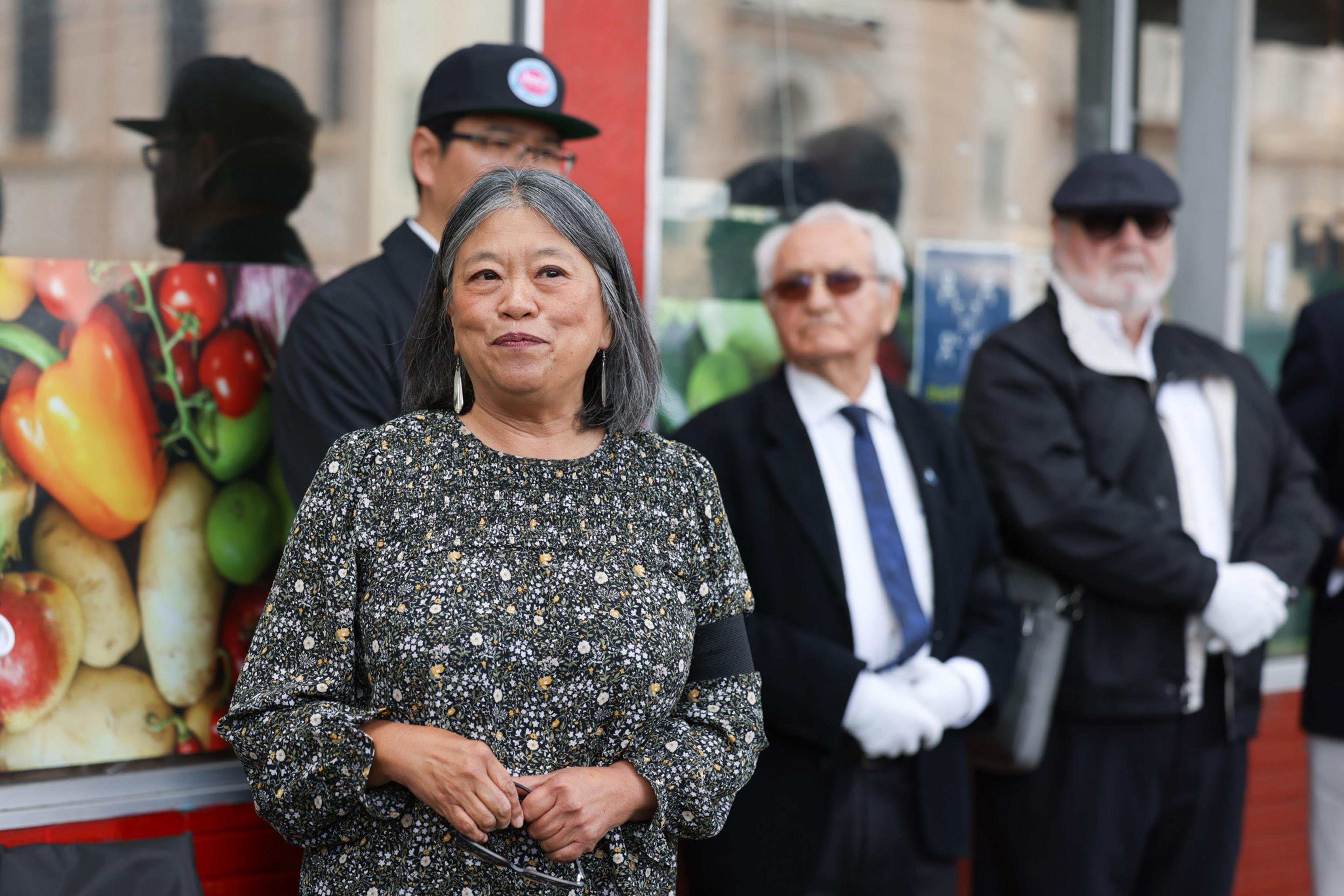 A woman in a patterned dress stands in front of three men dressed in formal black and white attire, with a background featuring colorful images of vegetables.