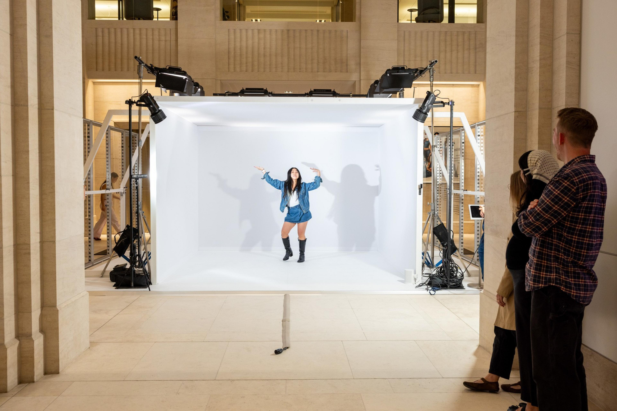 A woman in a blue jacket, shorts, and black boots poses inside a well-lit white photo booth while three people watch from outside.