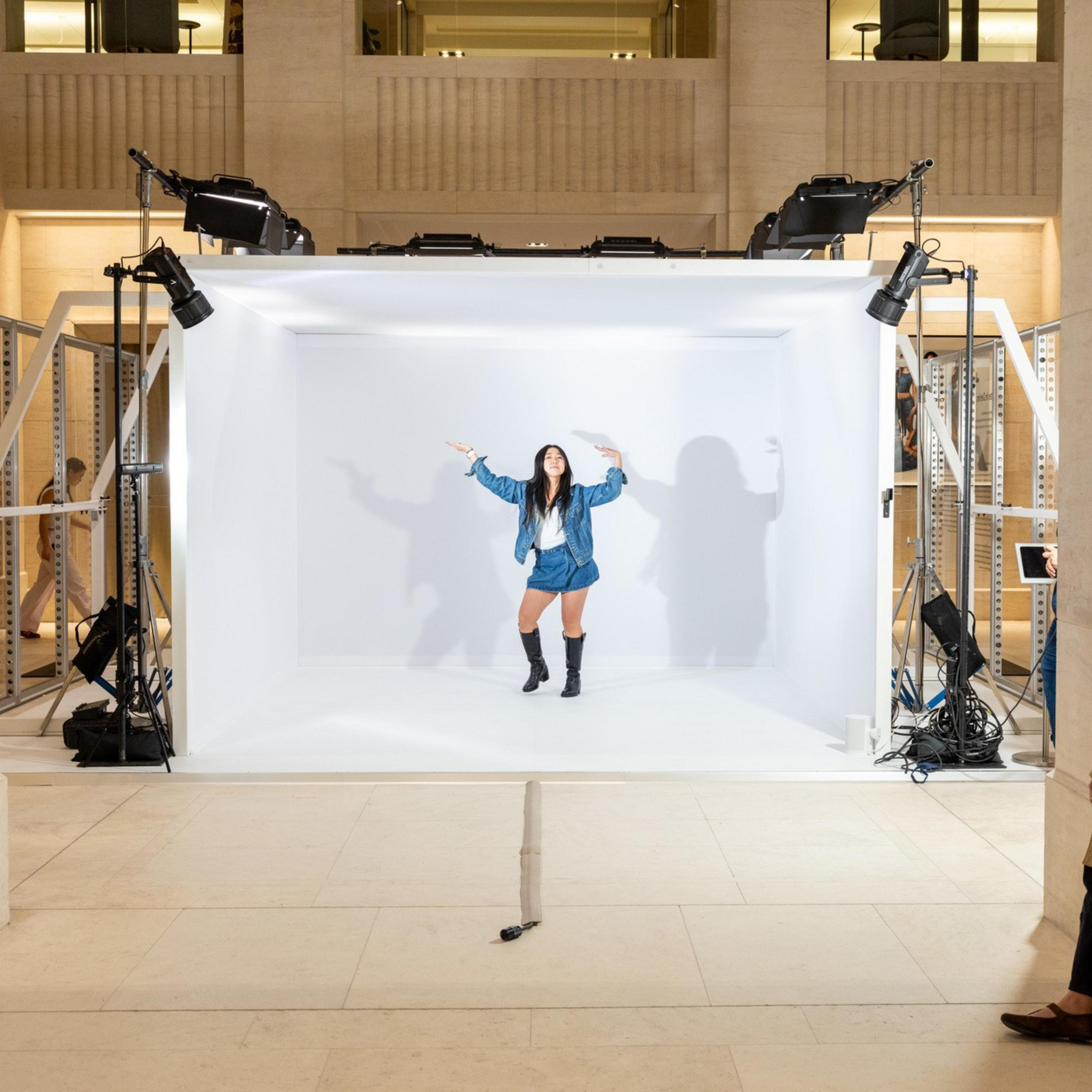 A woman in a blue jacket, shorts, and black boots poses inside a well-lit white photo booth while three people watch from outside.