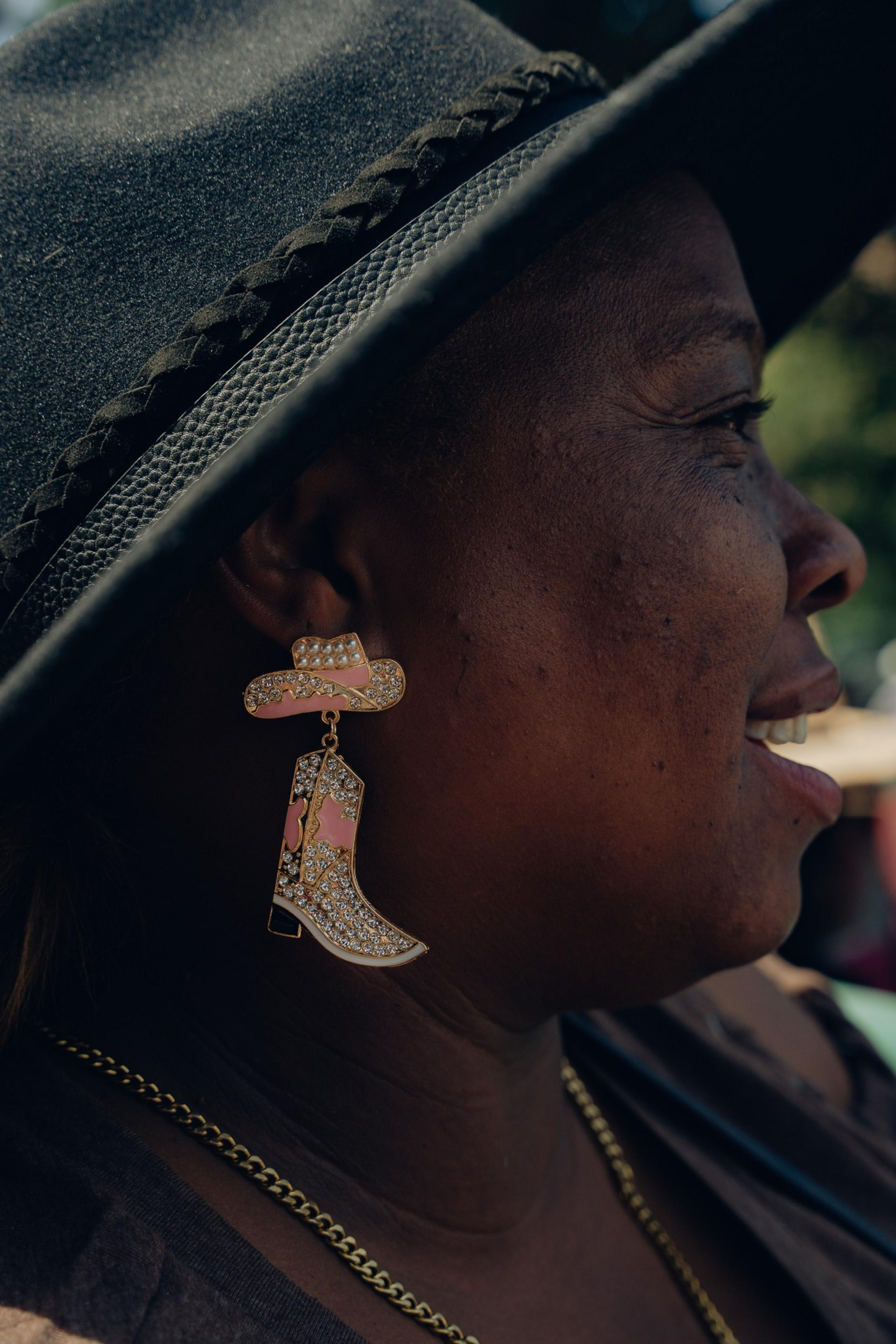 A person wearing a black hat with a braided band, a gold chain necklace, and an earring shaped like a rhinestone-decorated cowboy hat and boot set.