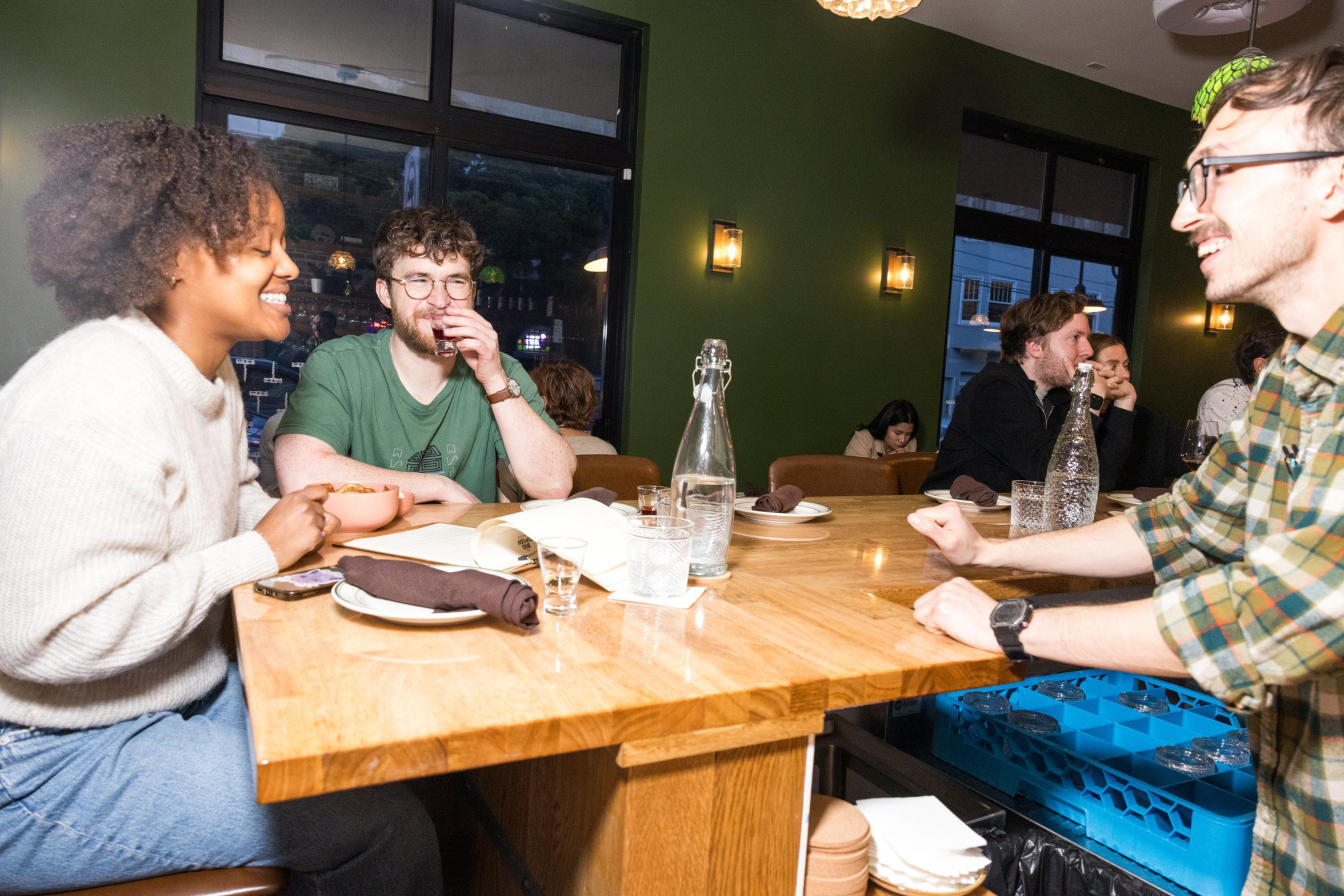 A group of people are sitting around a wooden table in a warmly-lit restaurant. They're smiling, talking, and appear to be enjoying their drinks.