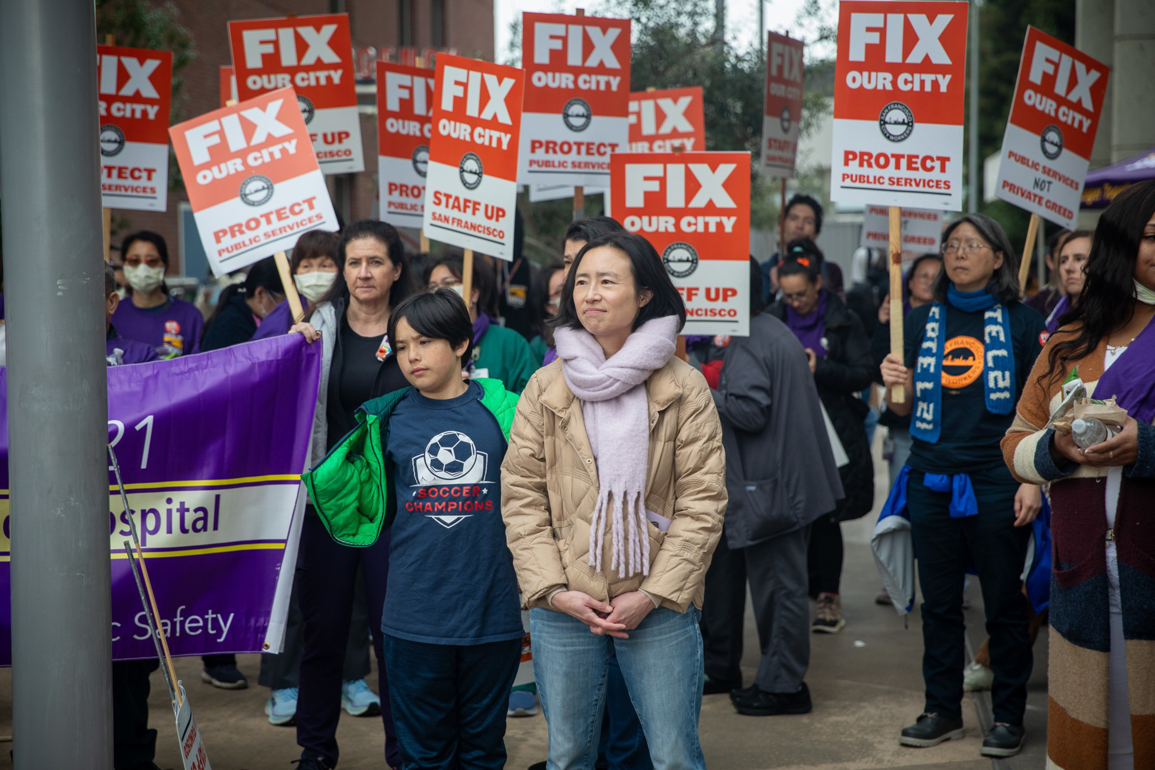 A crowd with &quot;FIX OUR CITY&quot; signs at a protest, a child in a soccer shirt looks forward.