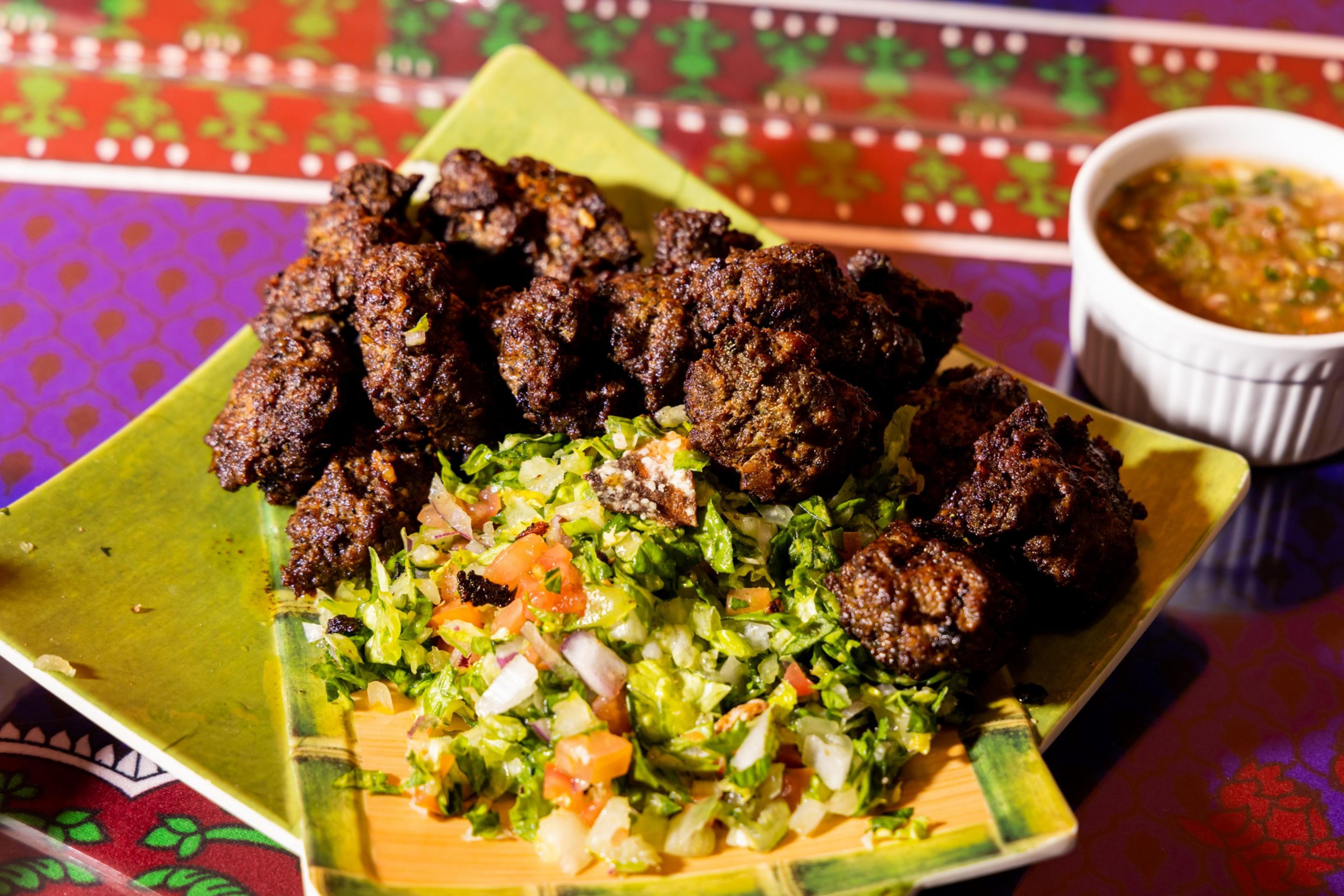 The image shows grilled meat pieces on a platter with chopped salad, including lettuce, tomatoes, and onions. A small cup of dipping sauce is to the side.