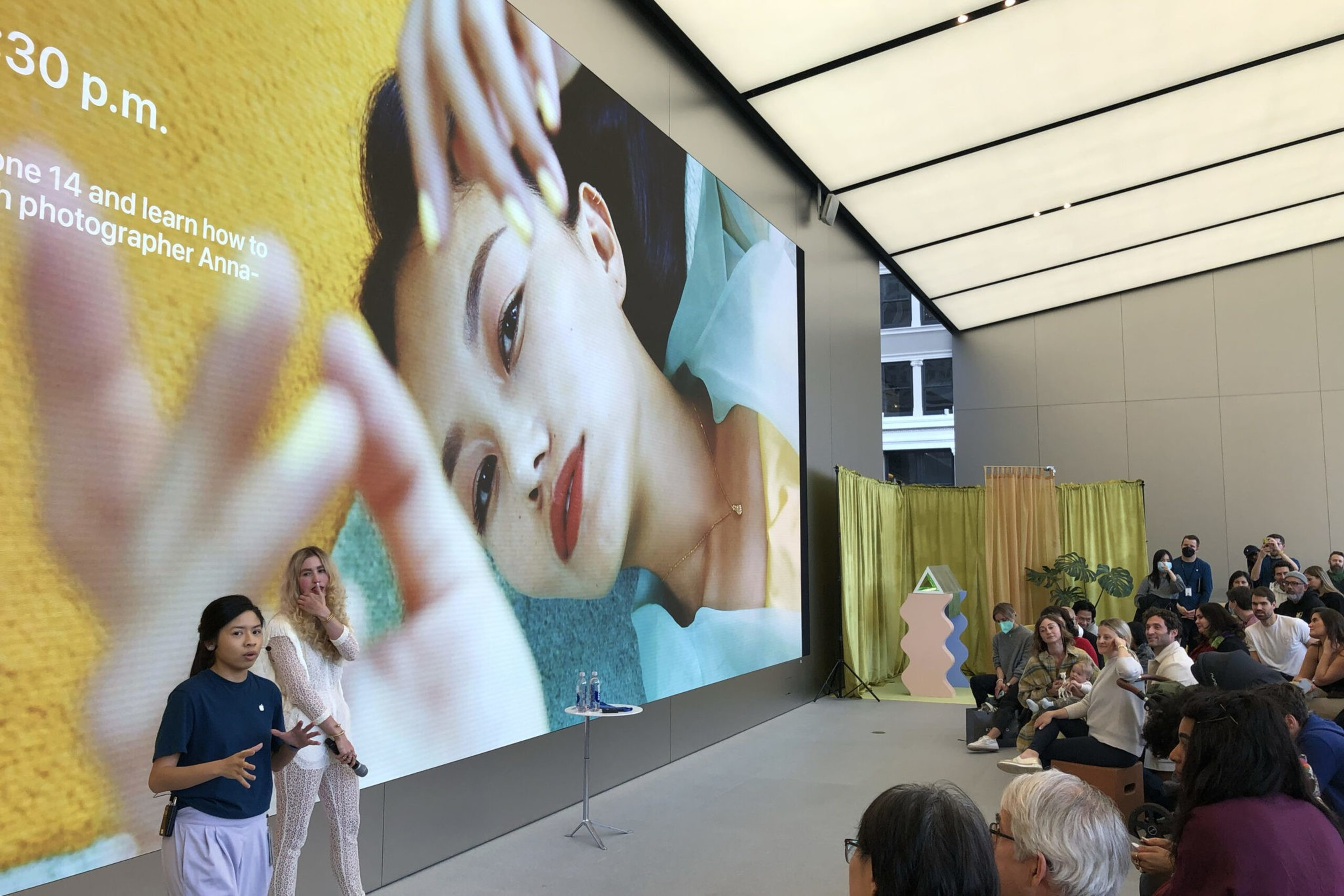 Two women stand presenting in front of a large screen displaying a close-up portrait of a woman with red lipstick and a teal background, while an audience watches.
