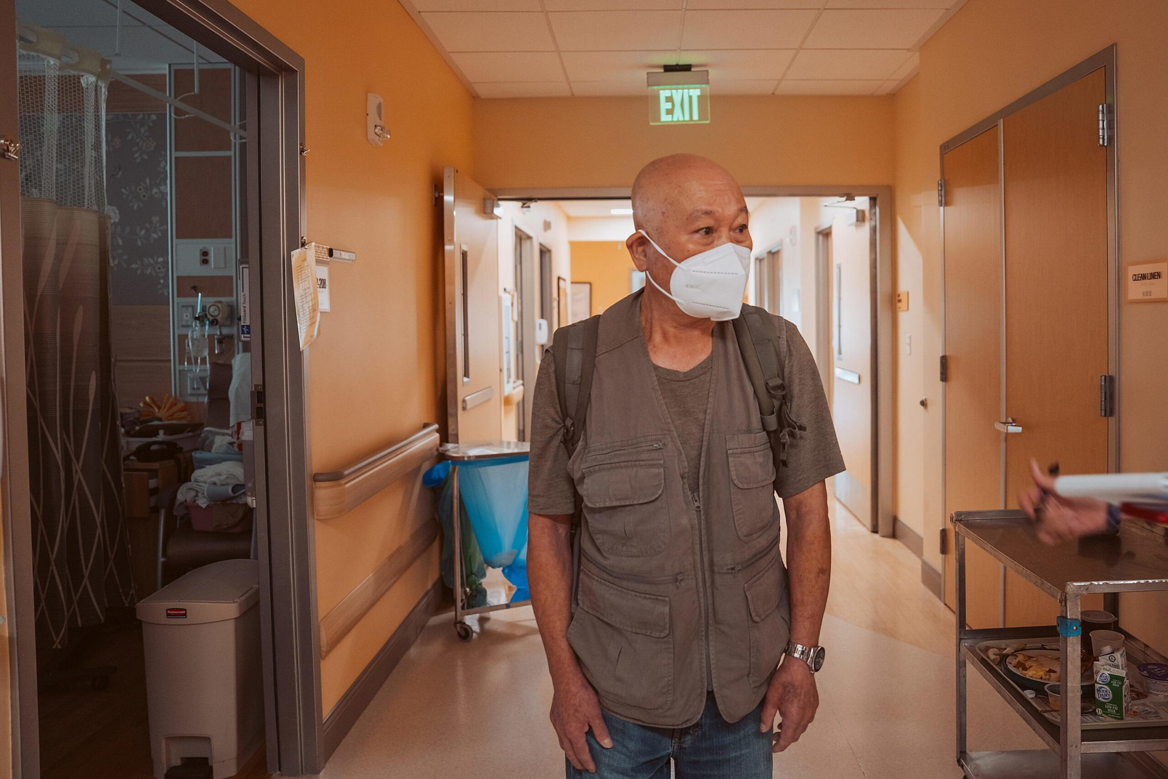 An older man wearing a face mask and backpack stands in a hospital hallway with orange walls and an exit sign overhead.