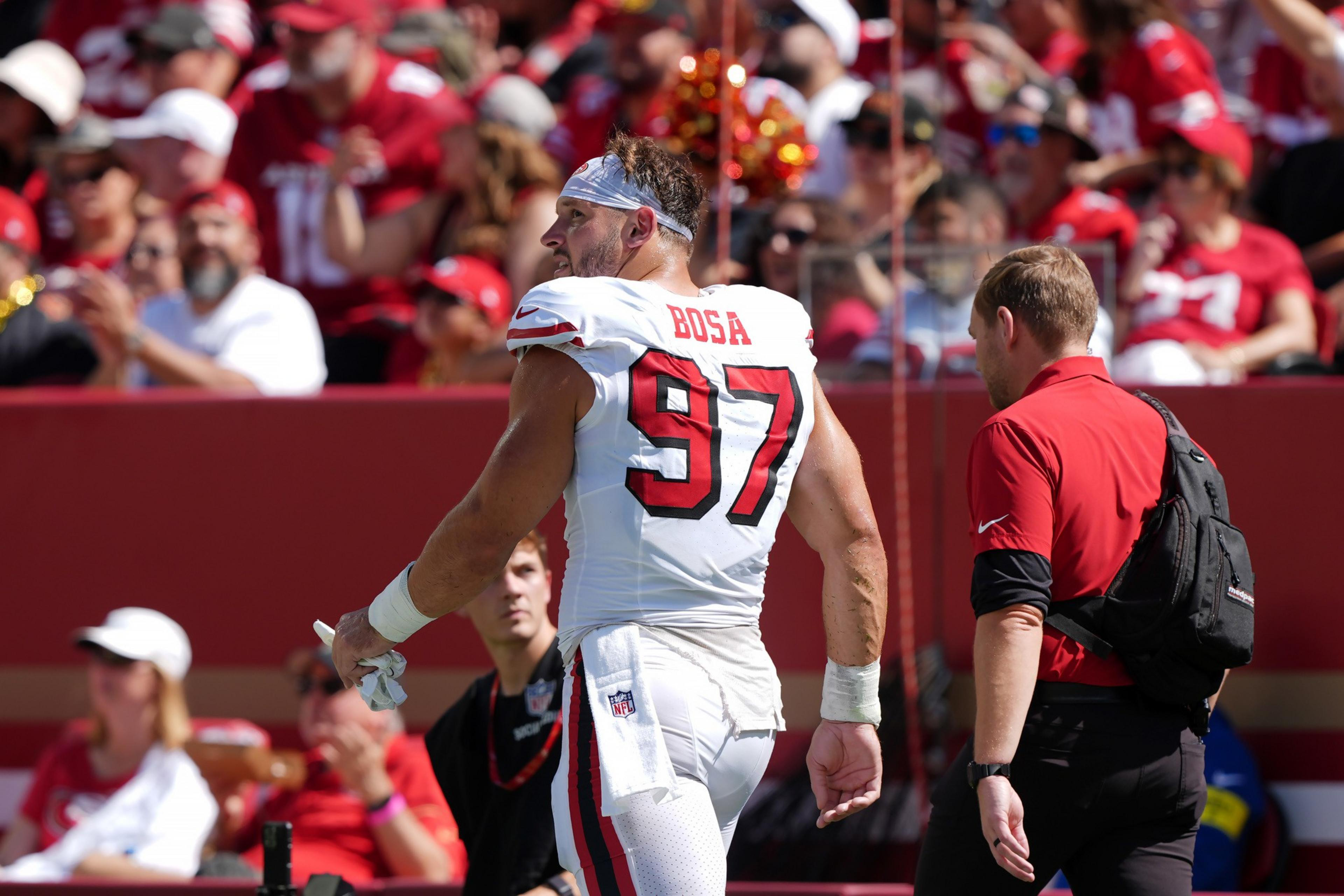 A football player wearing a white jersey with “BOSA 97" on the back stands on the field next to a man in a red shirt and black backpack.