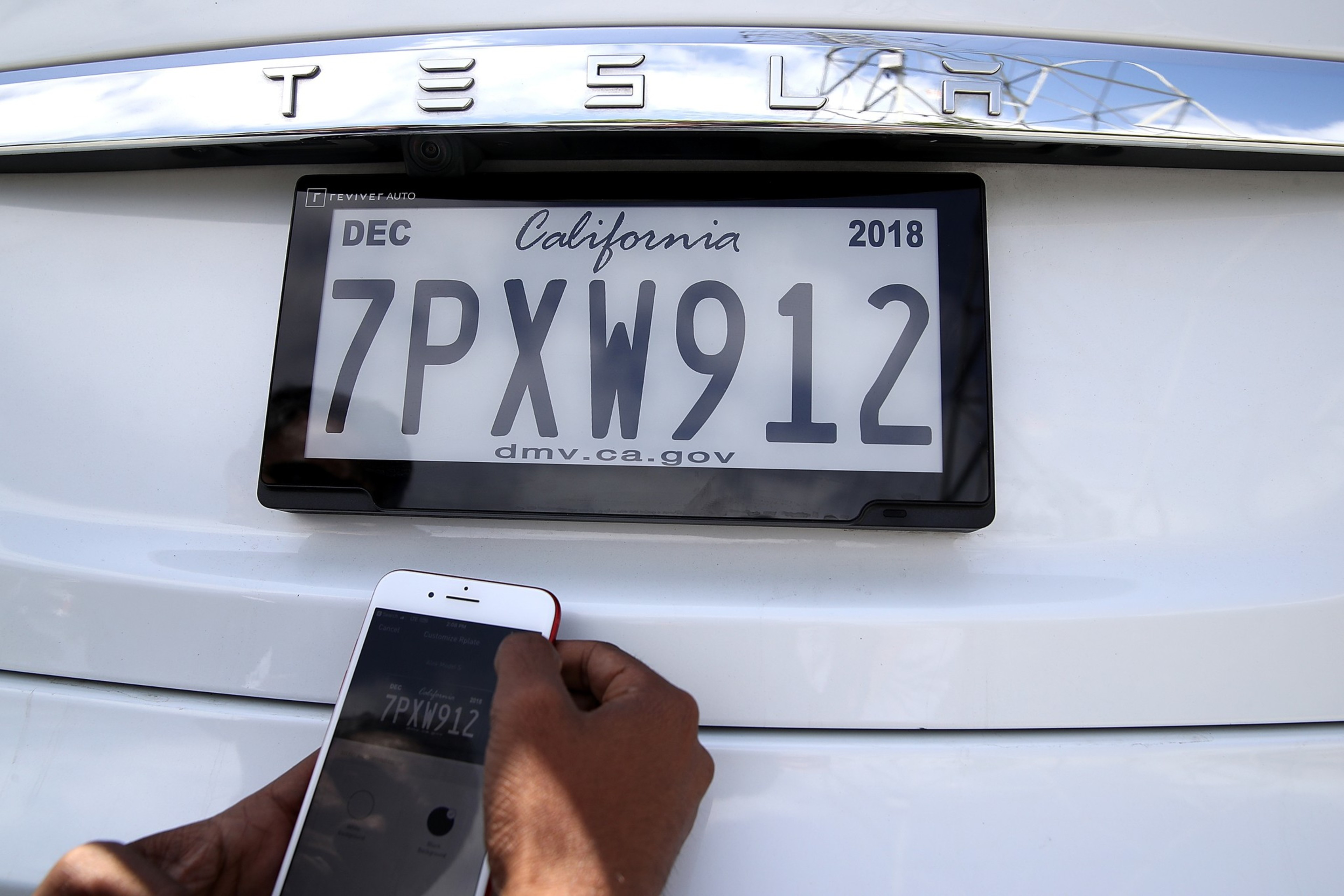 A person is holding a smartphone, matching a digital car plate on a white Tesla with "TESLA" logo above.