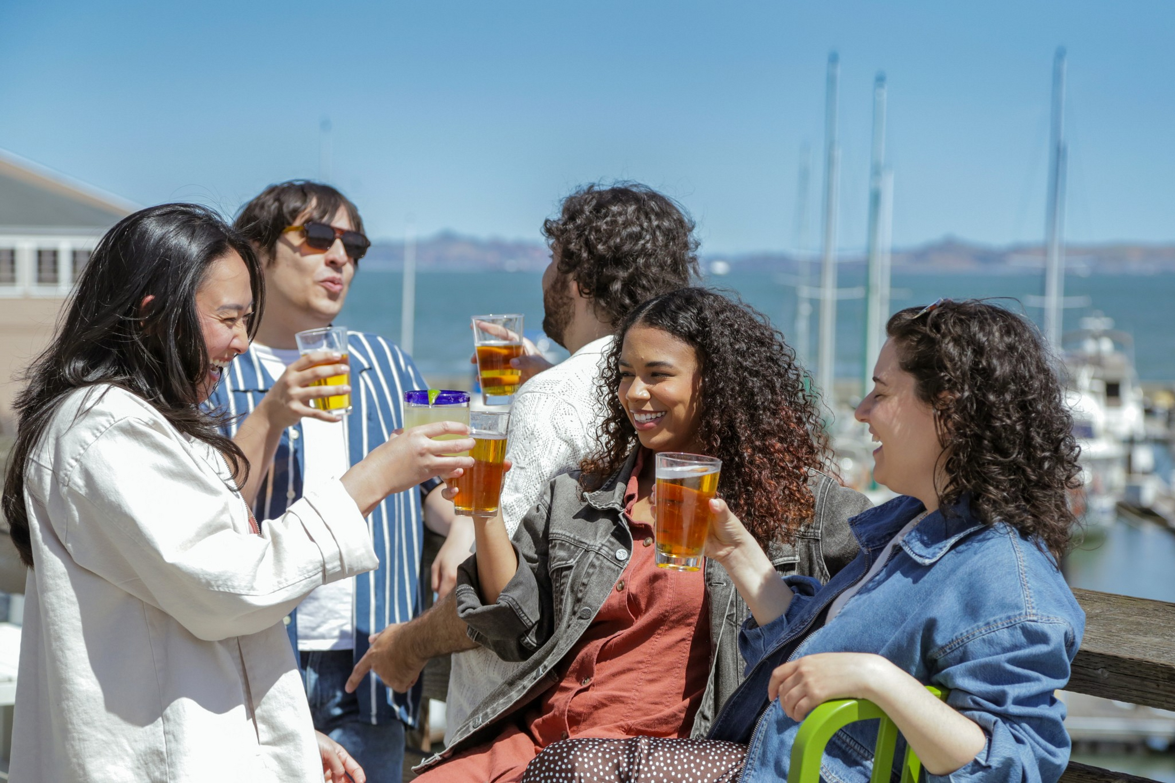 Five friends laugh and toast with glasses of beer on a sunny dockside with sailboats and water in the background.