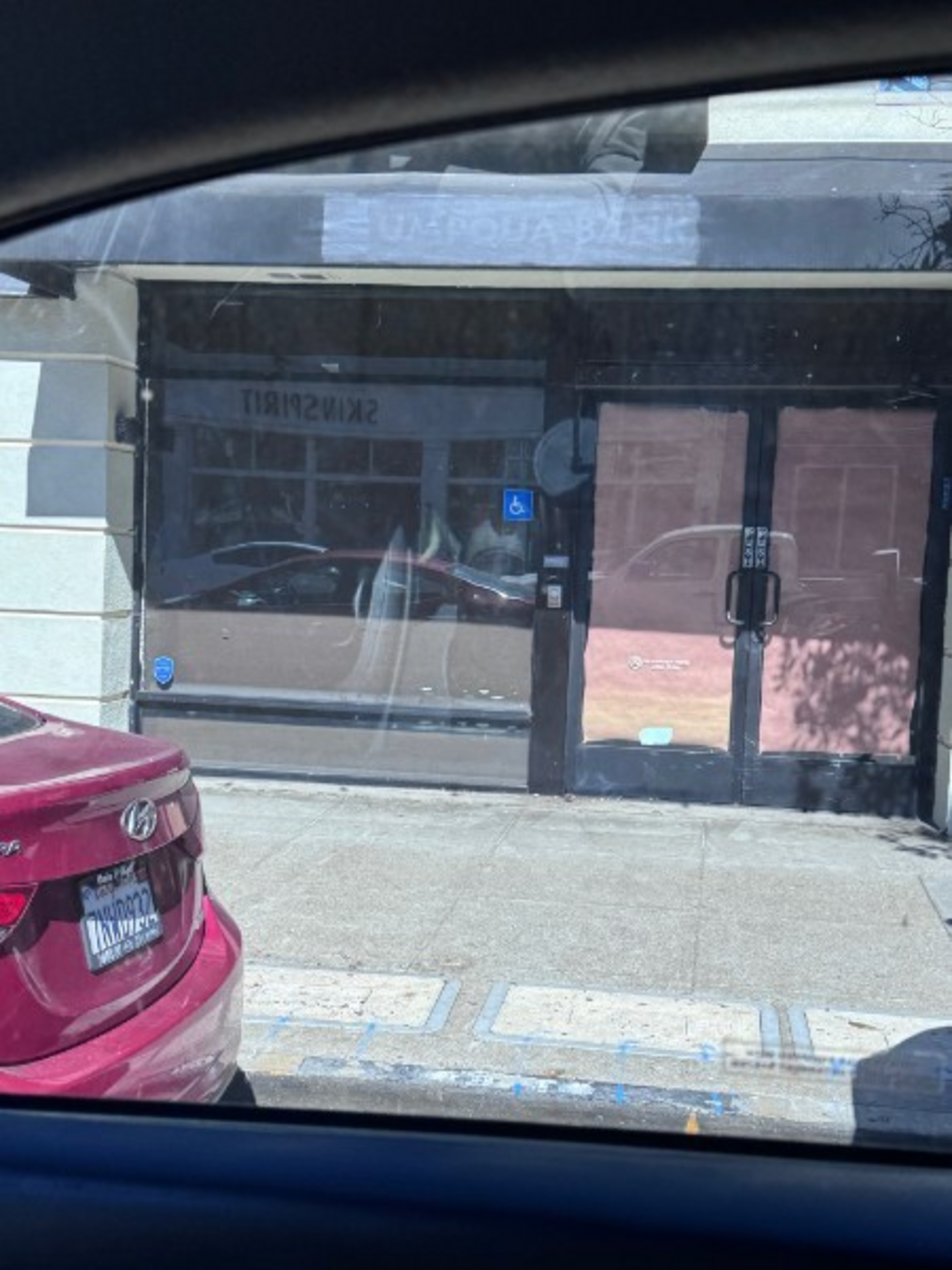 A vacant storefront with covered windows and double glass doors sits next to a red car parked in a handicapped parking spot.