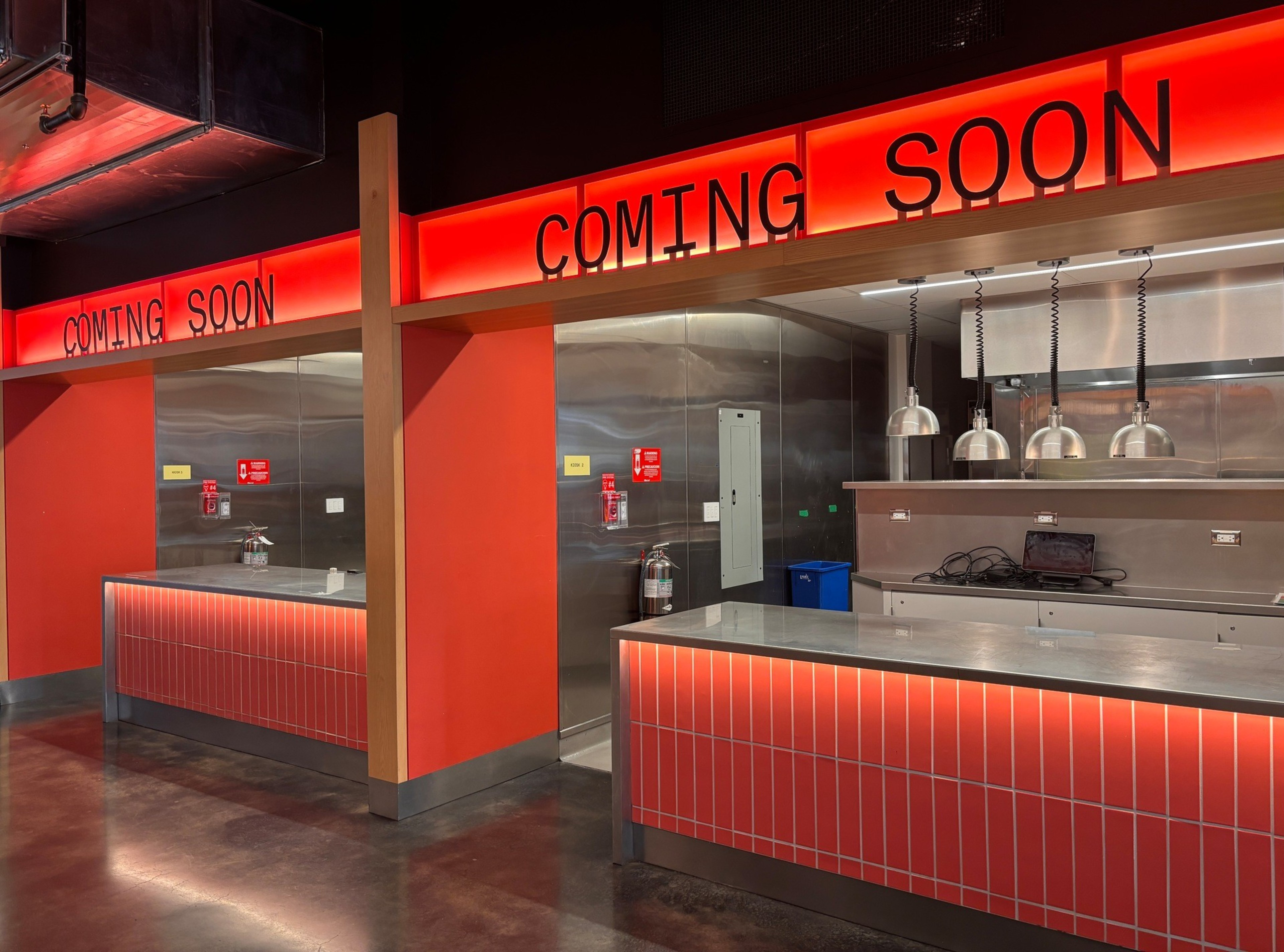 Two empty, modern service counters with red tiled fronts and stainless steel surfaces display "COMING SOON" in black letters on bright red backlit signs above.