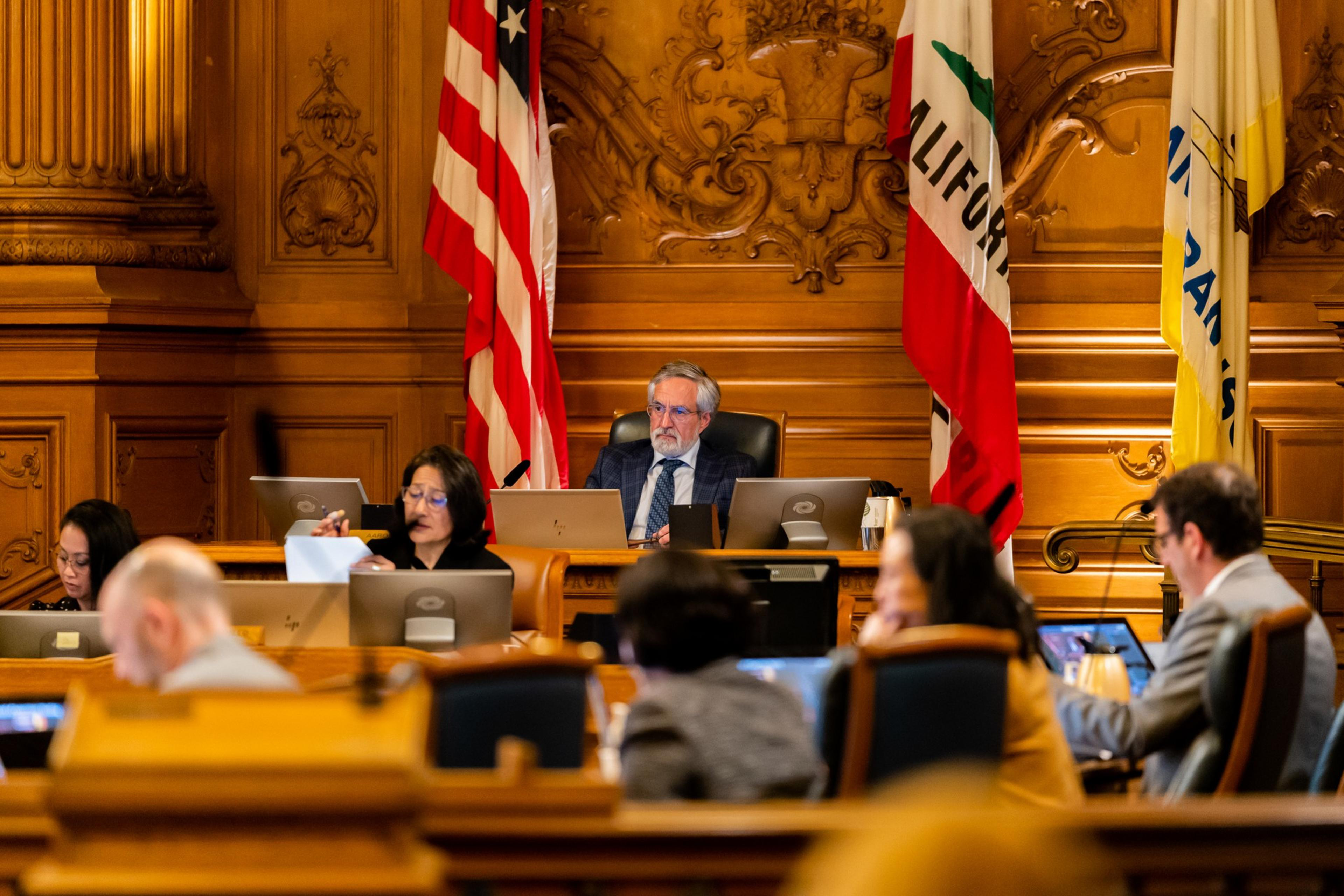 A formal meeting with officials seated at desks, the American and California flags behind a central man in a suit, wood-paneled walls, and laptops in use.