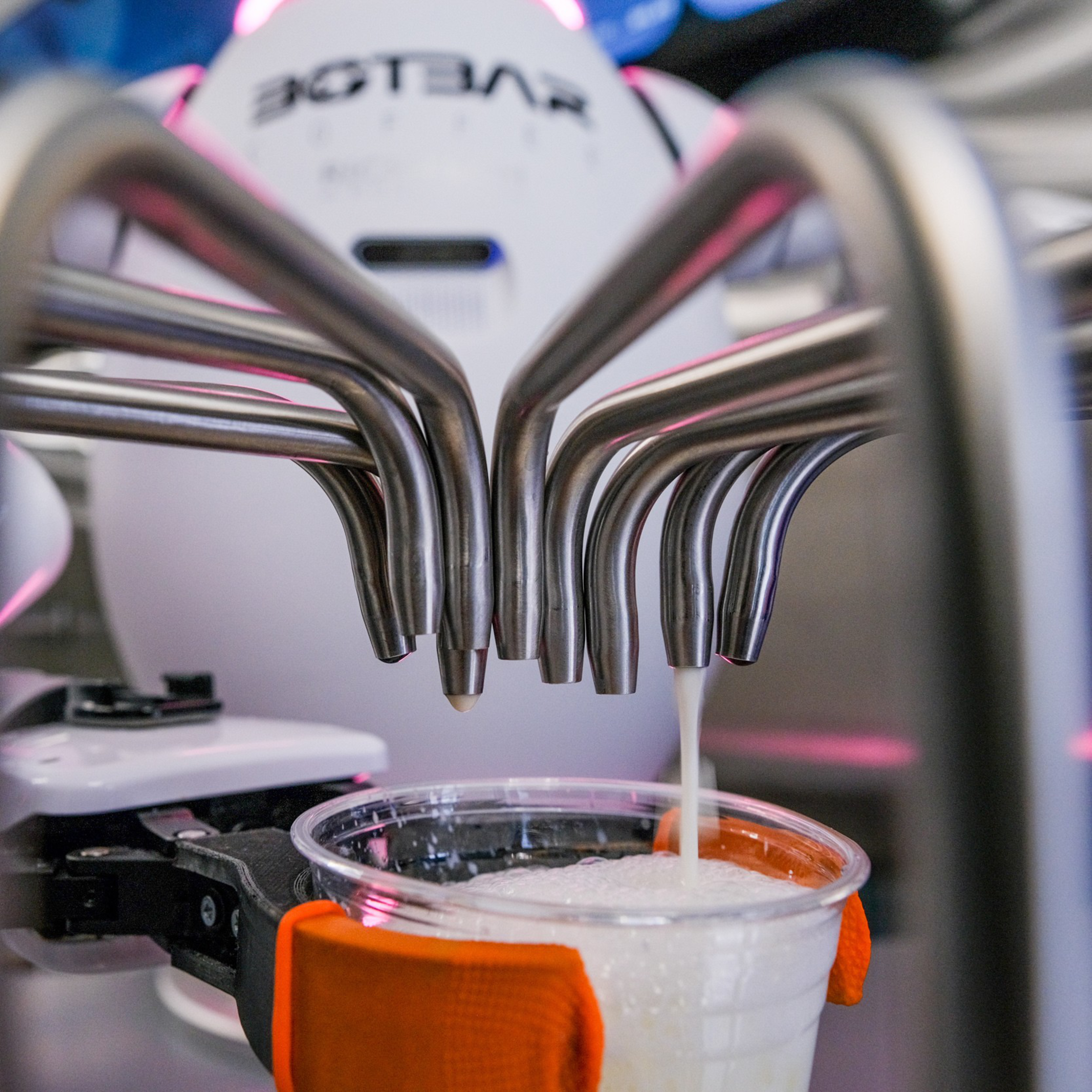 A robotic arm holds a plastic cup under several metallic nozzles, which are dispensing a foamy liquid into the cup.