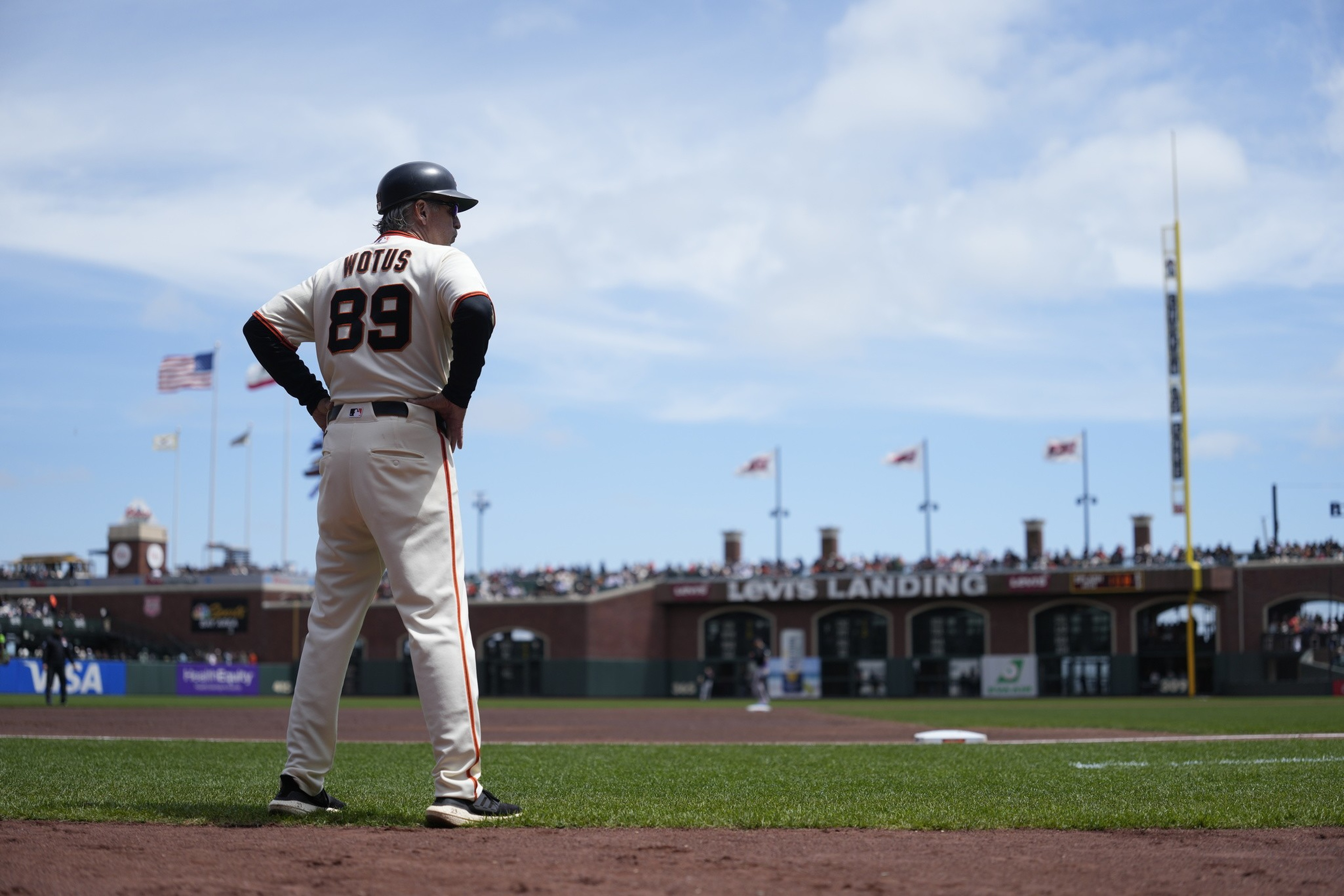 A baseball coach wearing a "NOTUS 89" jersey stands near the field's edge, facing the game with a crowd and flags visible in the background.