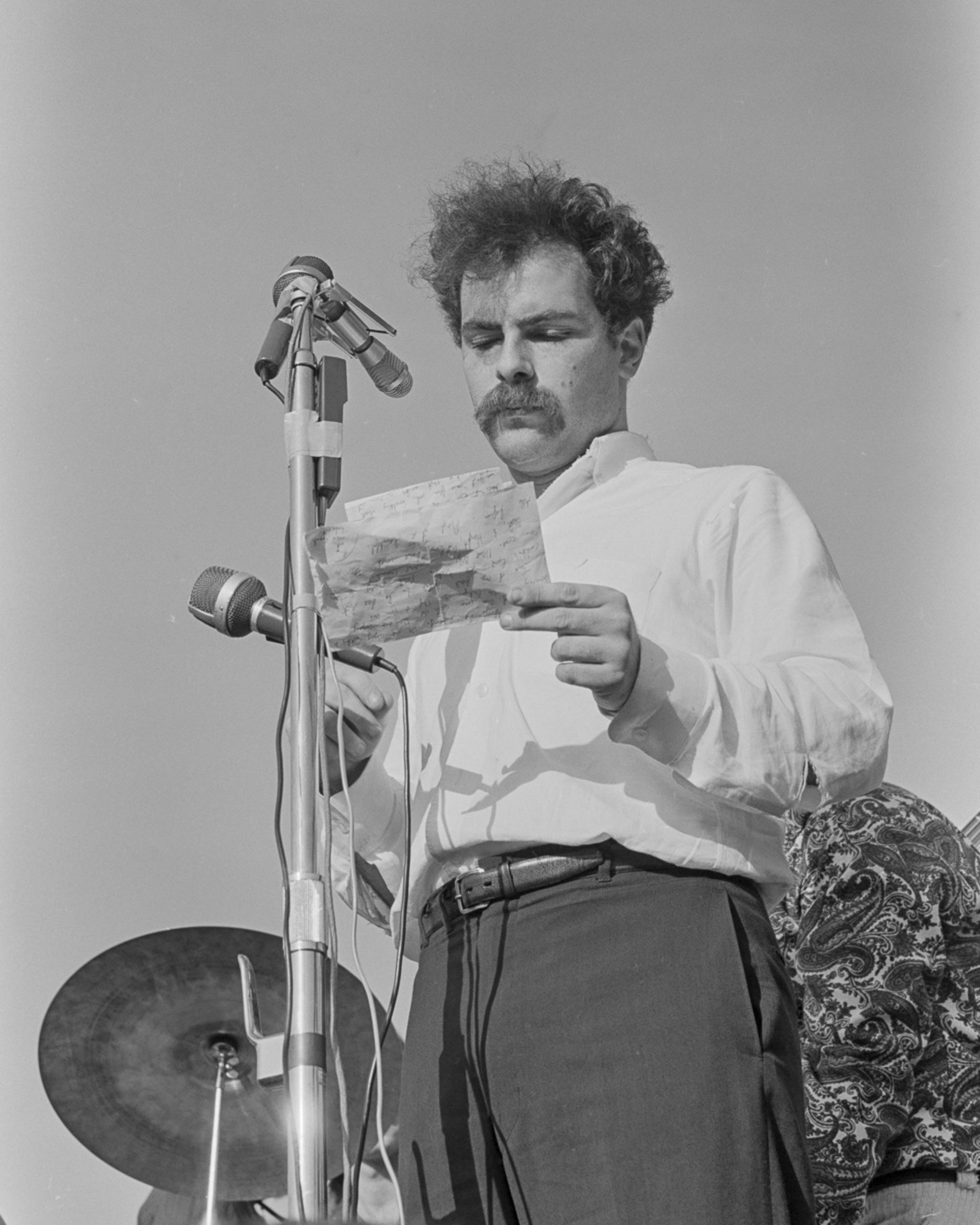 A man with curly hair and a mustache reads from a paper in front of microphones. He's wearing a white shirt, standing near a musical instrument outdoors.