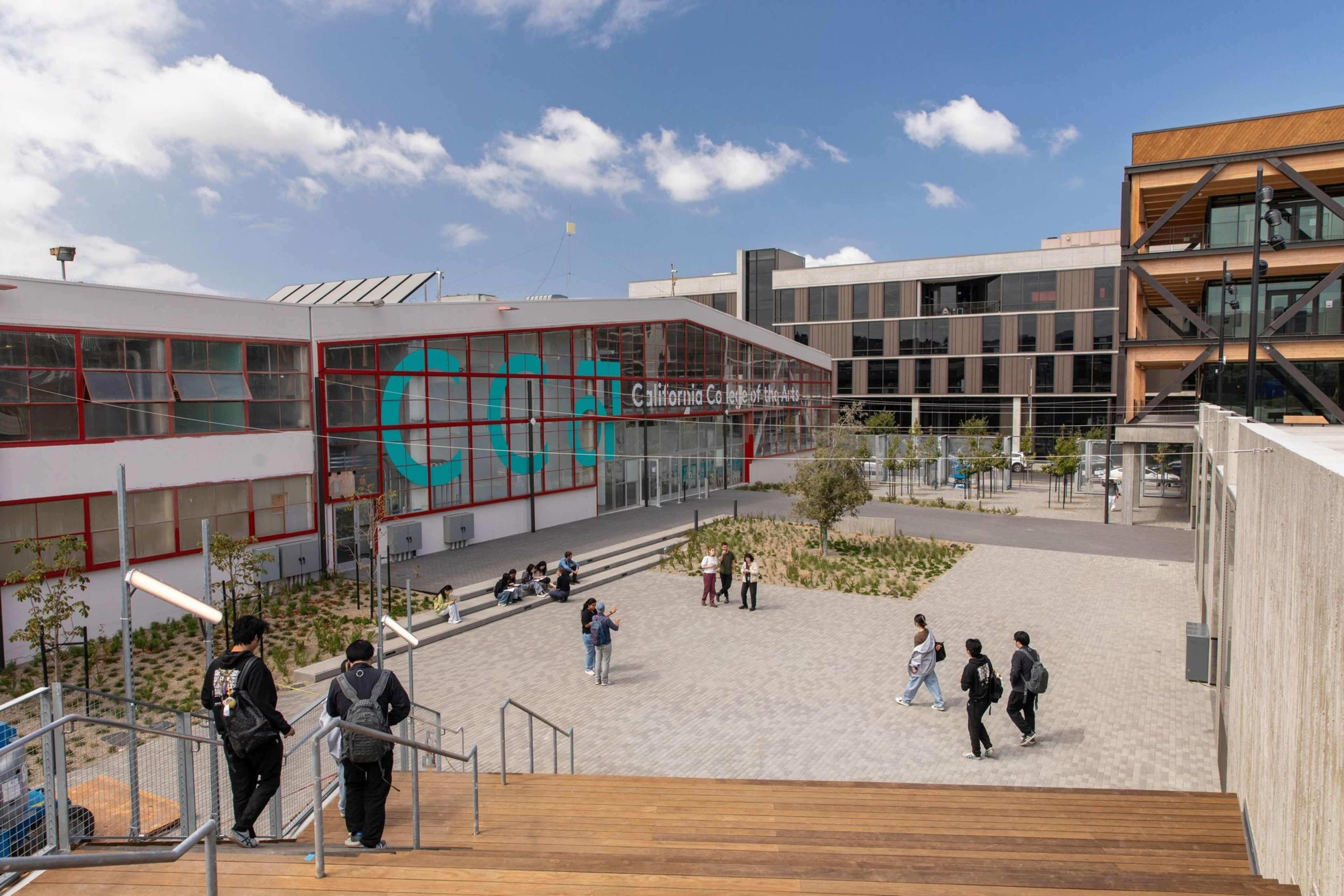 A modern campus setting features a building with large &quot;CCA&quot; letters and students walking across a spacious, tree-lined courtyard under a partly cloudy sky.