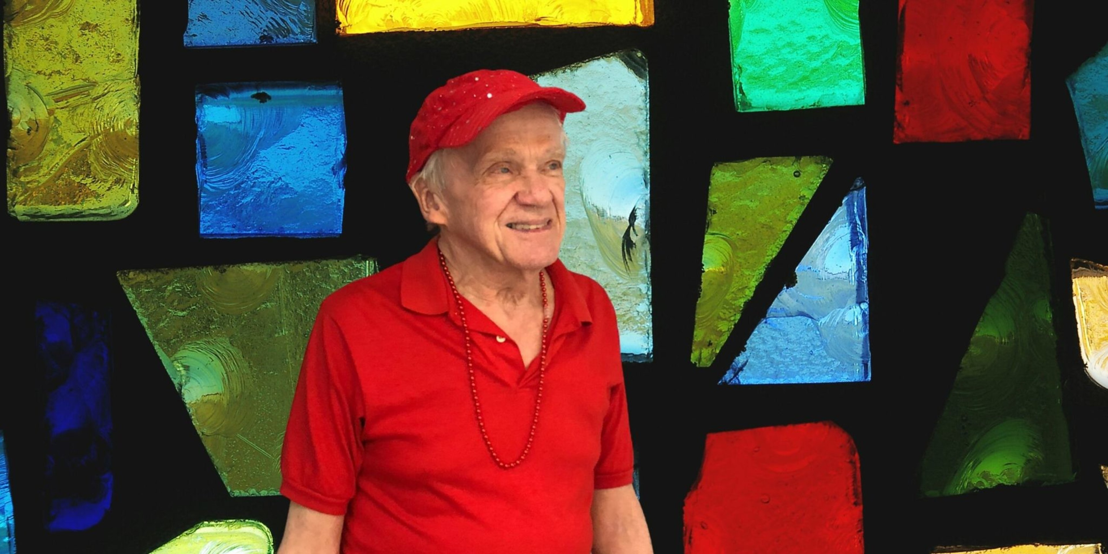 An elderly man in a red shirt, hat, and necklace stands smiling in front of a colorful stained glass window with blue, green, red, and yellow panes.