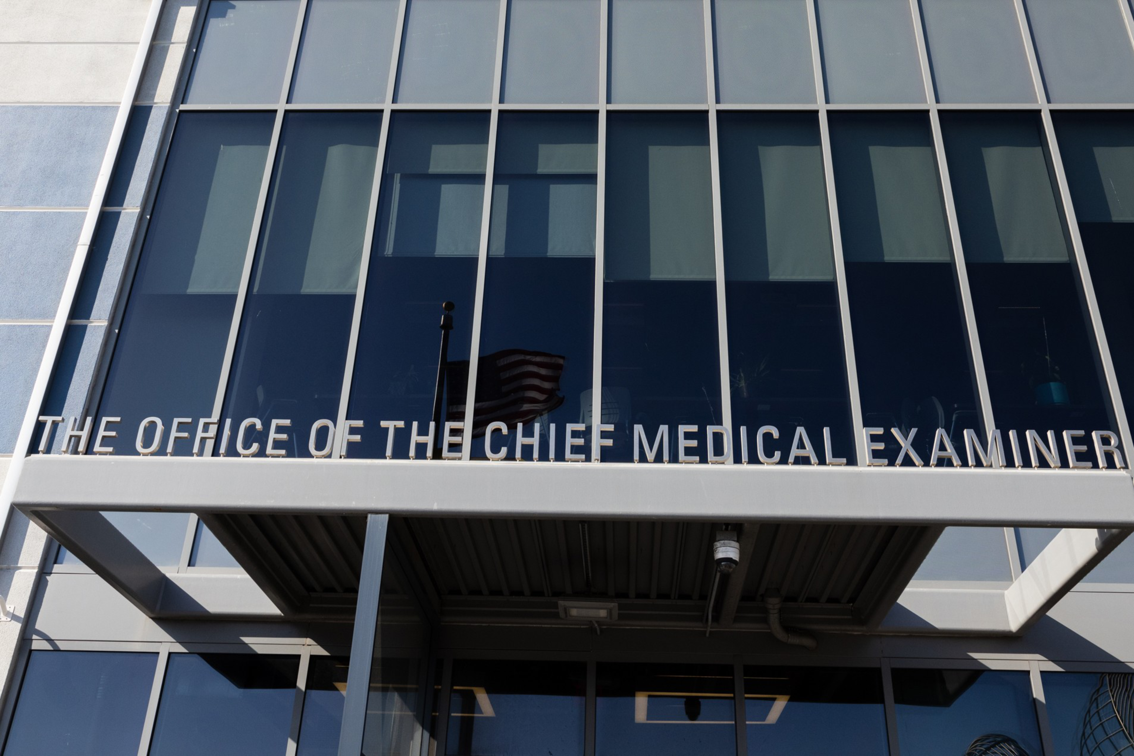 A glass building facade displays the sign "The Office of the Chief Medical Examiner" above an entrance with a white overhang.