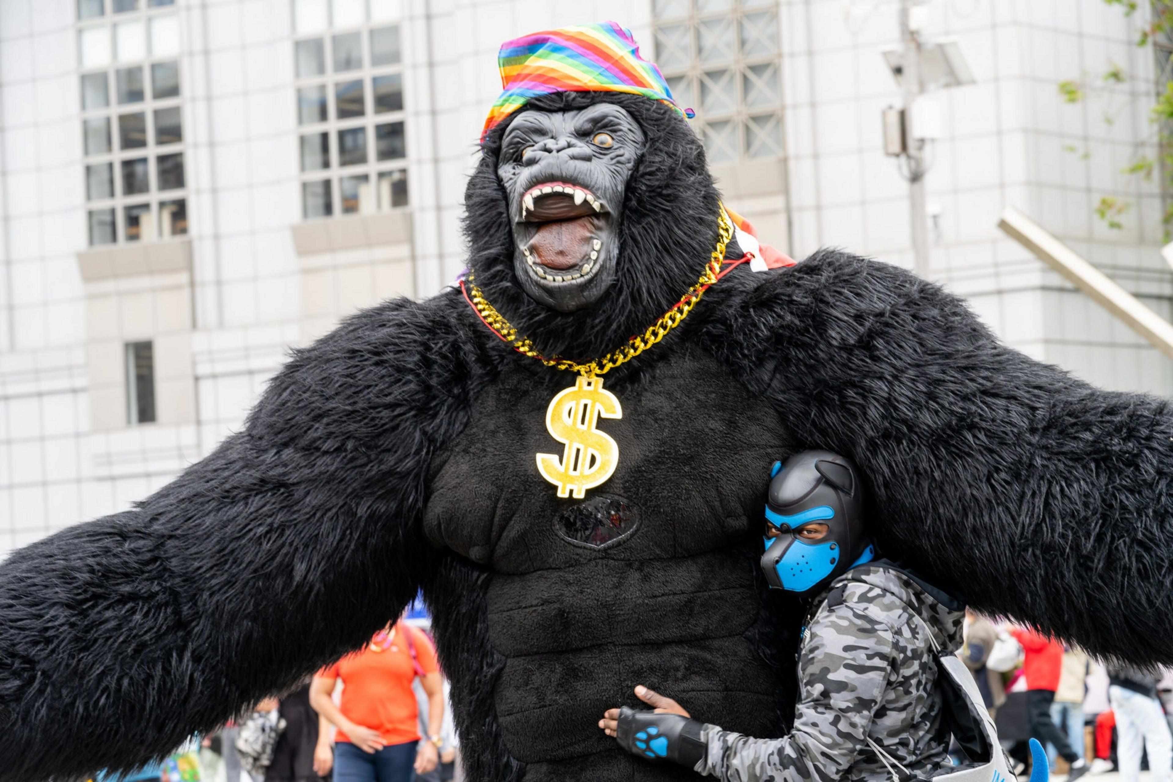 A person in a large gorilla costume wearing a colorful hat and gold dollar sign necklace is being hugged by someone in a blue dog mask and camo jacket.