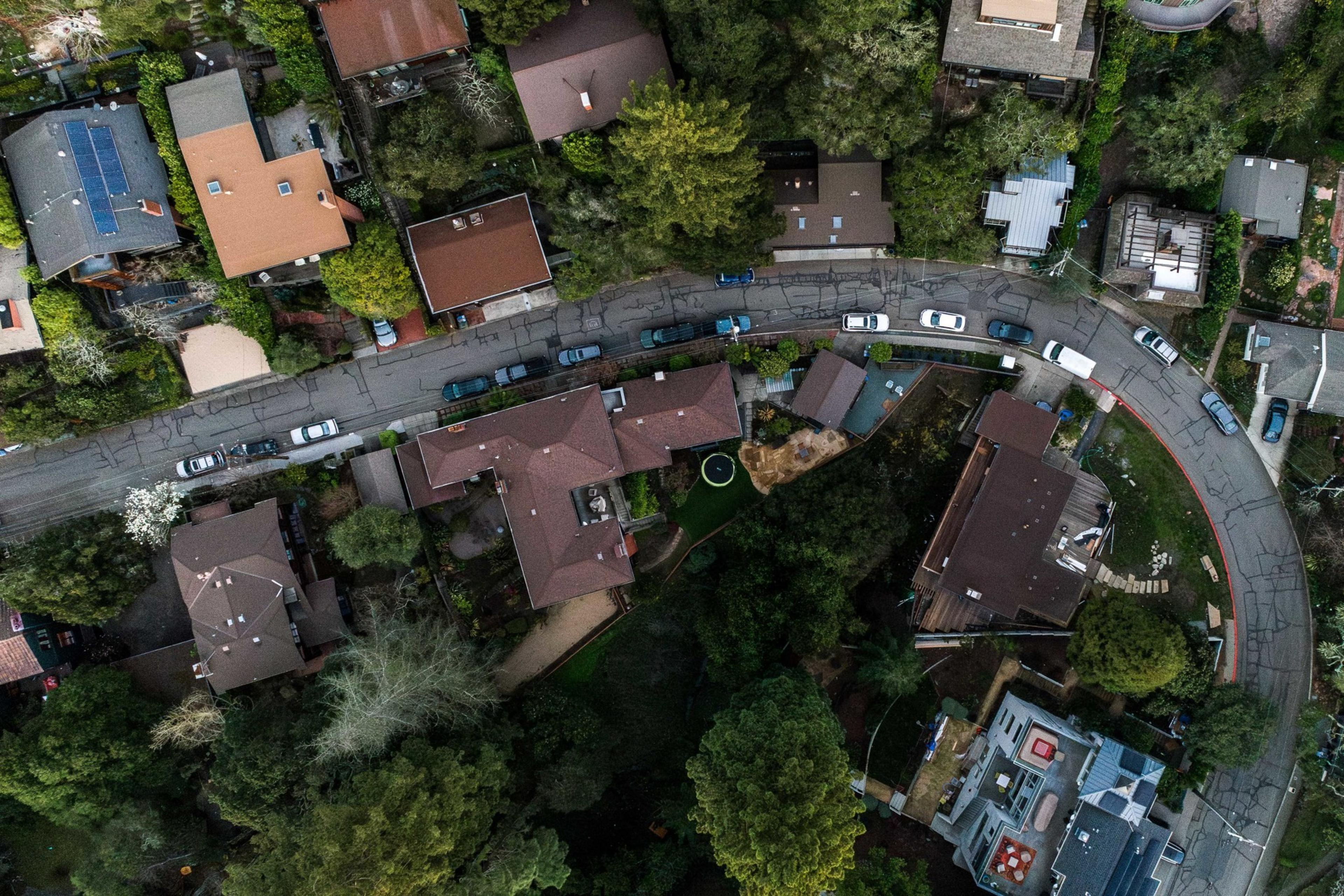 The image shows an aerial view of a suburban neighborhood with houses, trees, and parked cars lining a curved street, surrounded by lush greenery.