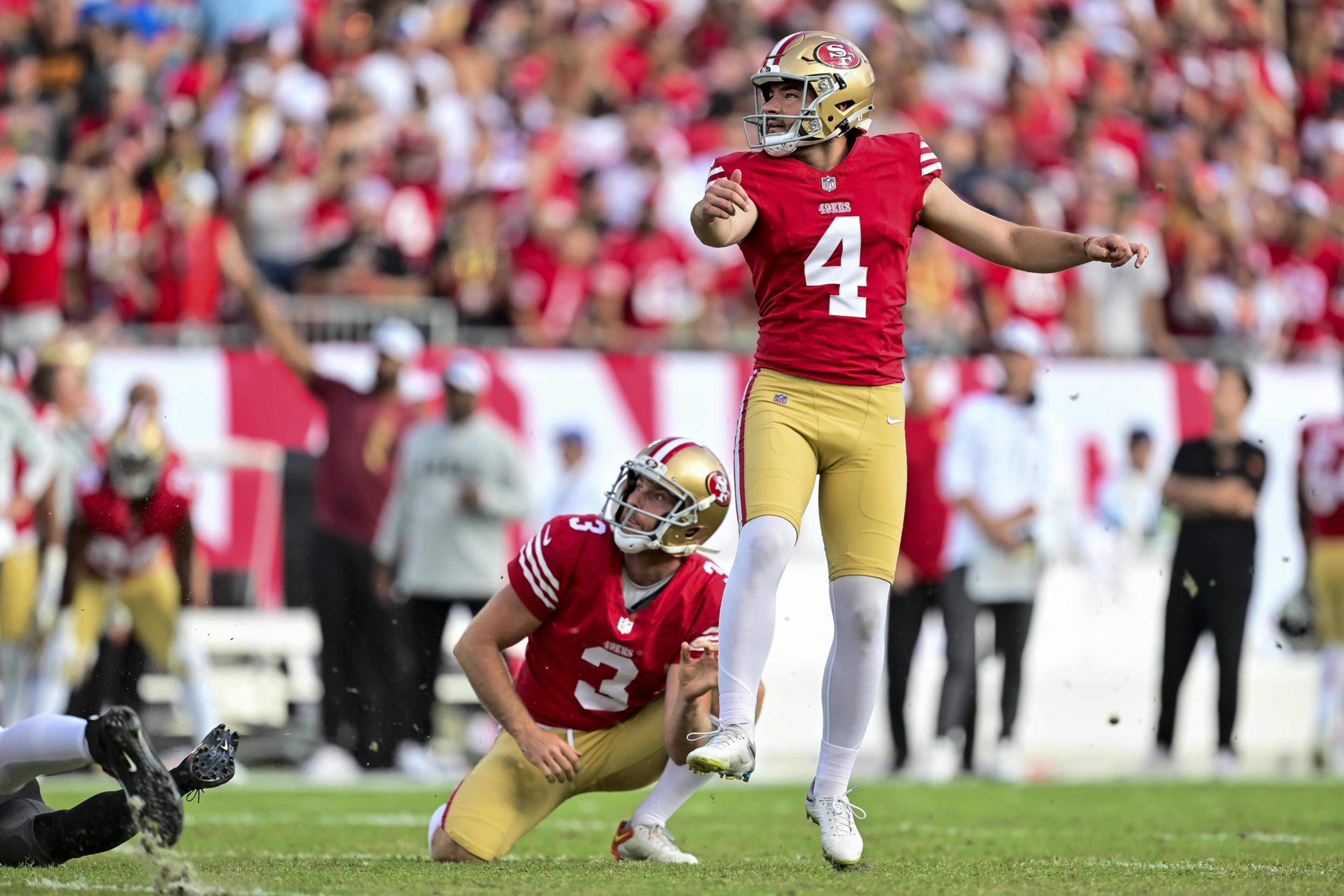 A football player in a red and gold uniform, number 4, is kicking a ball with a teammate, number 3, holding it on a field surrounded by a cheering crowd.
