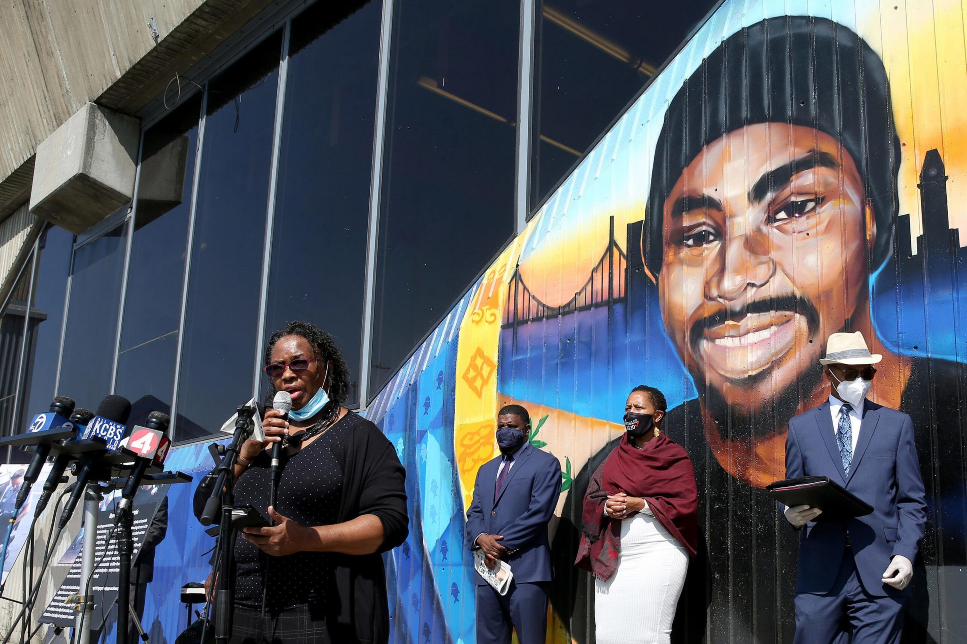 A group of people stand near microphones in front of a large mural of a smiling man wearing a beanie, with a cityscape and bridge in the background.
