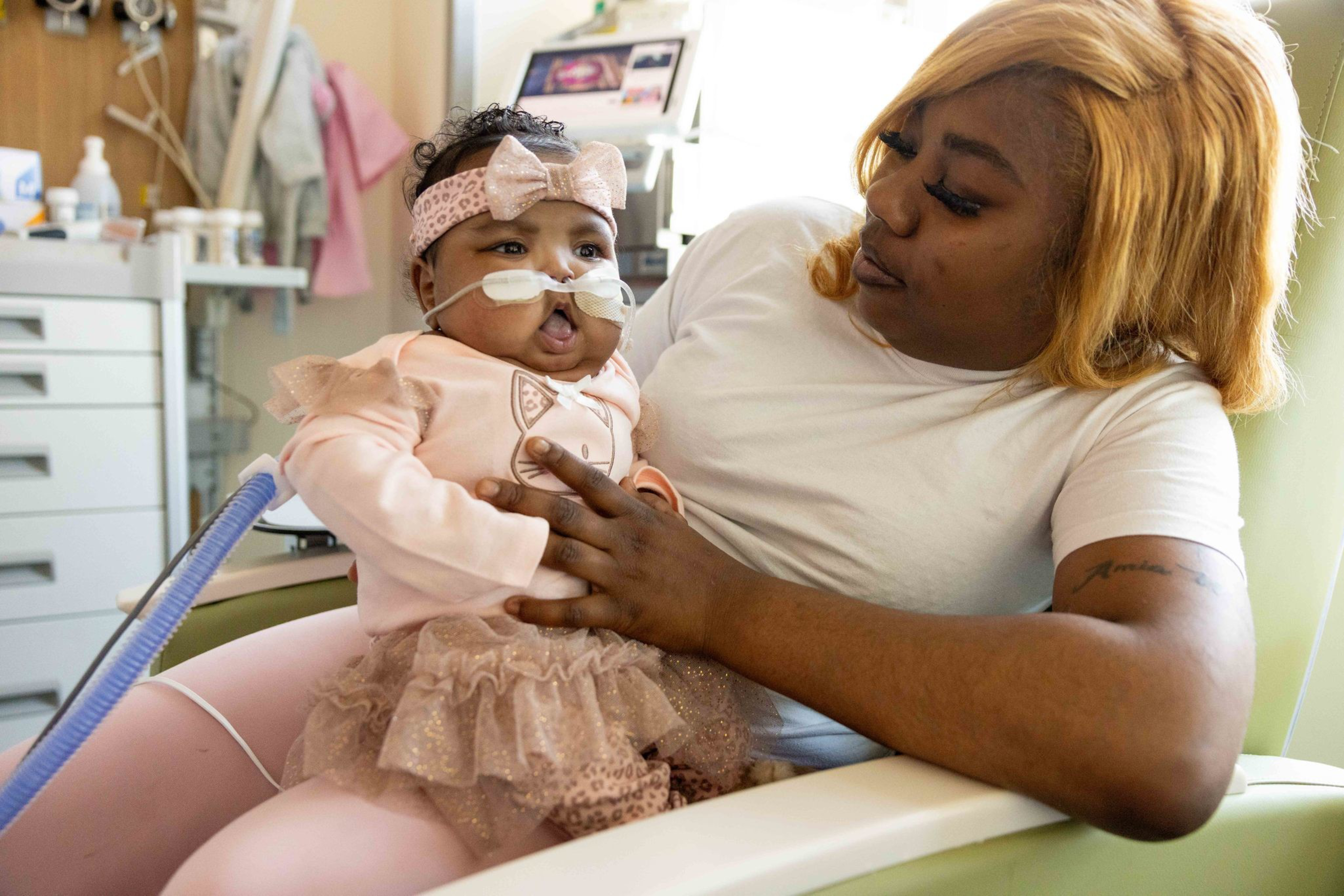 A woman with blonde hair gently holds a baby wearing a pink outfit with a headband and a nasal oxygen tube in a medical setting.