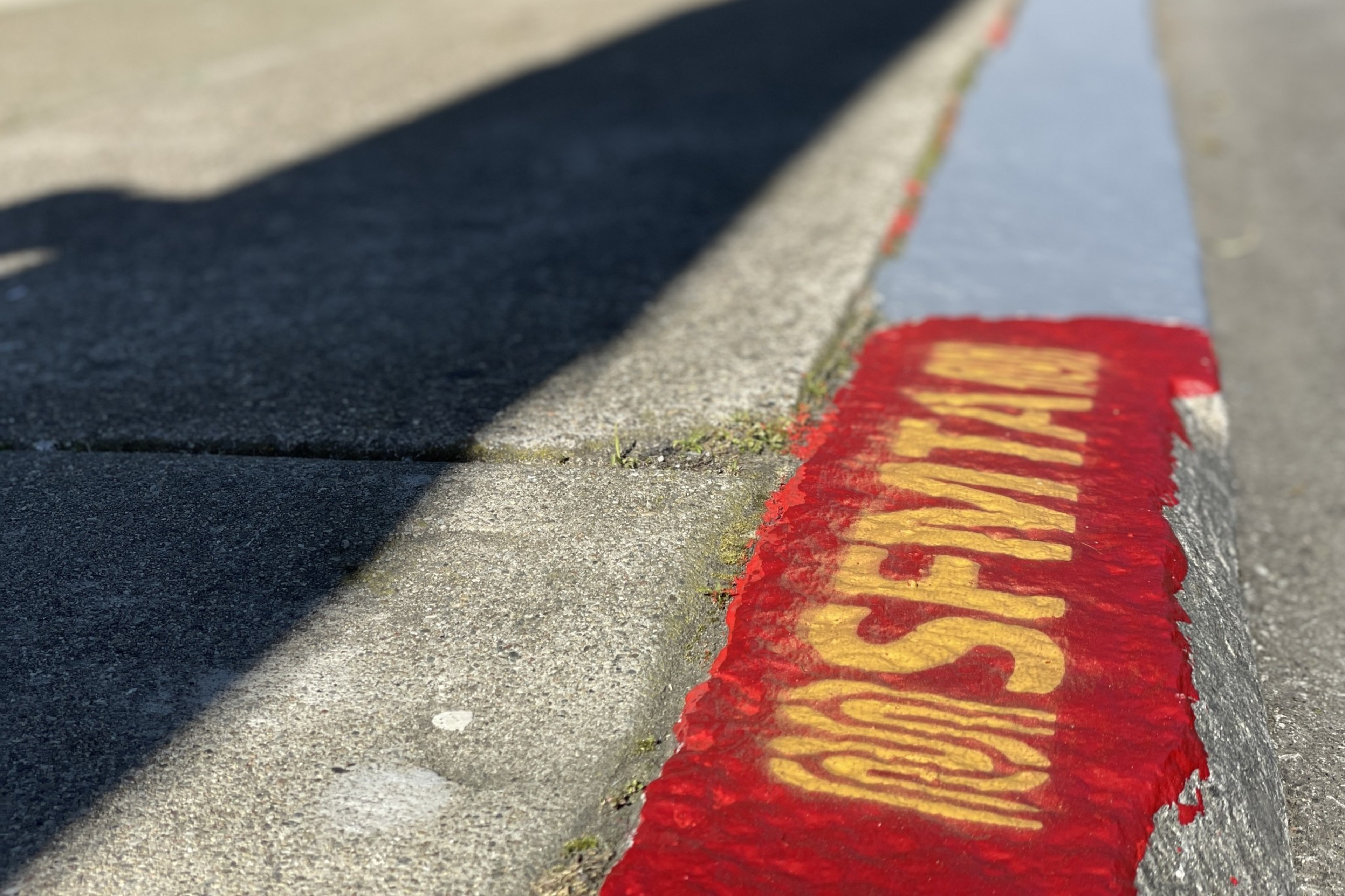 A sidewalk curb with "SFMTA" stenciled in yellow on red paint, casting shadows on the pavement in bright sunlight.