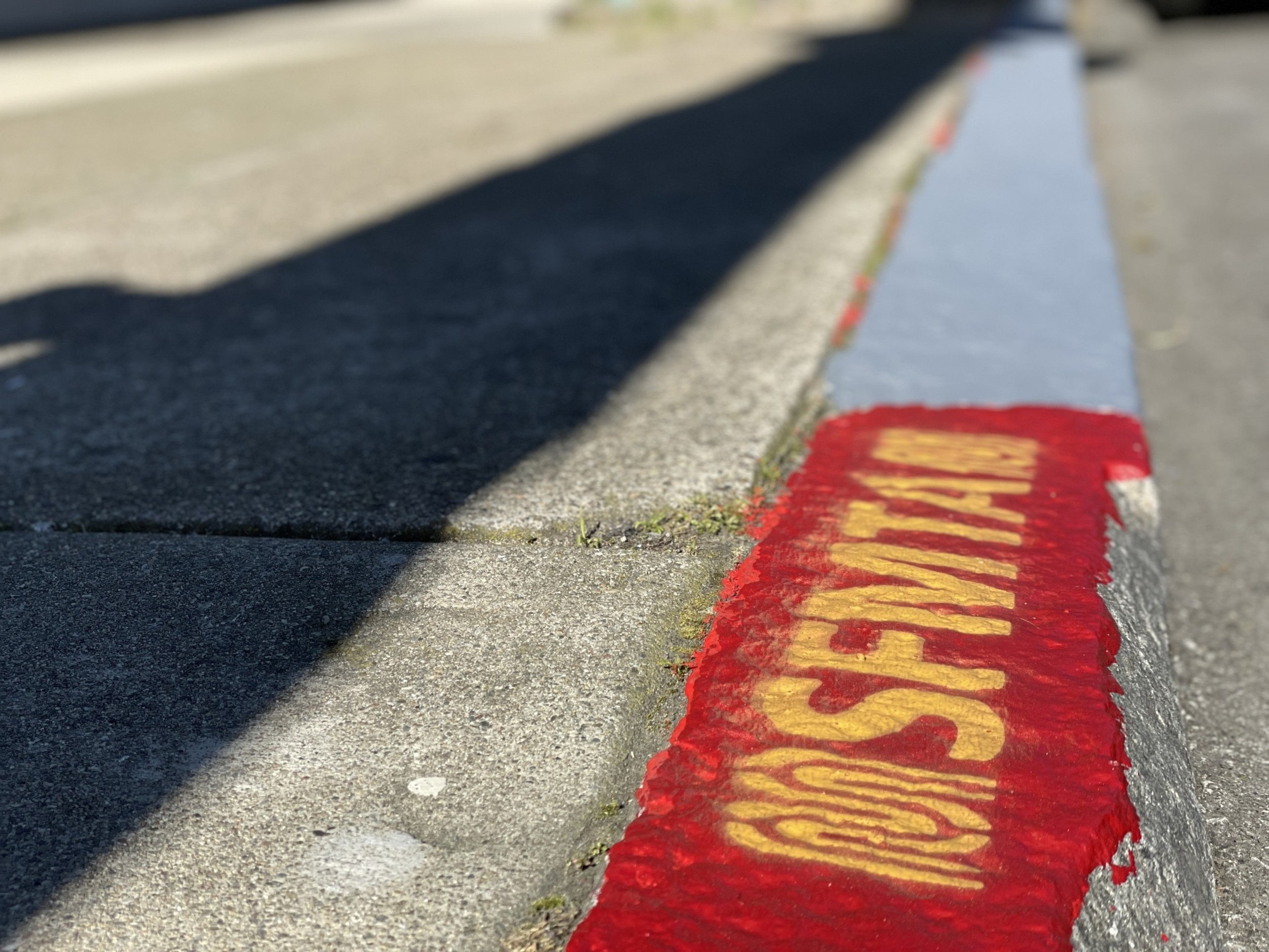 A sidewalk curb with "SFMTA" stenciled in yellow on red paint, casting shadows on the pavement in bright sunlight.