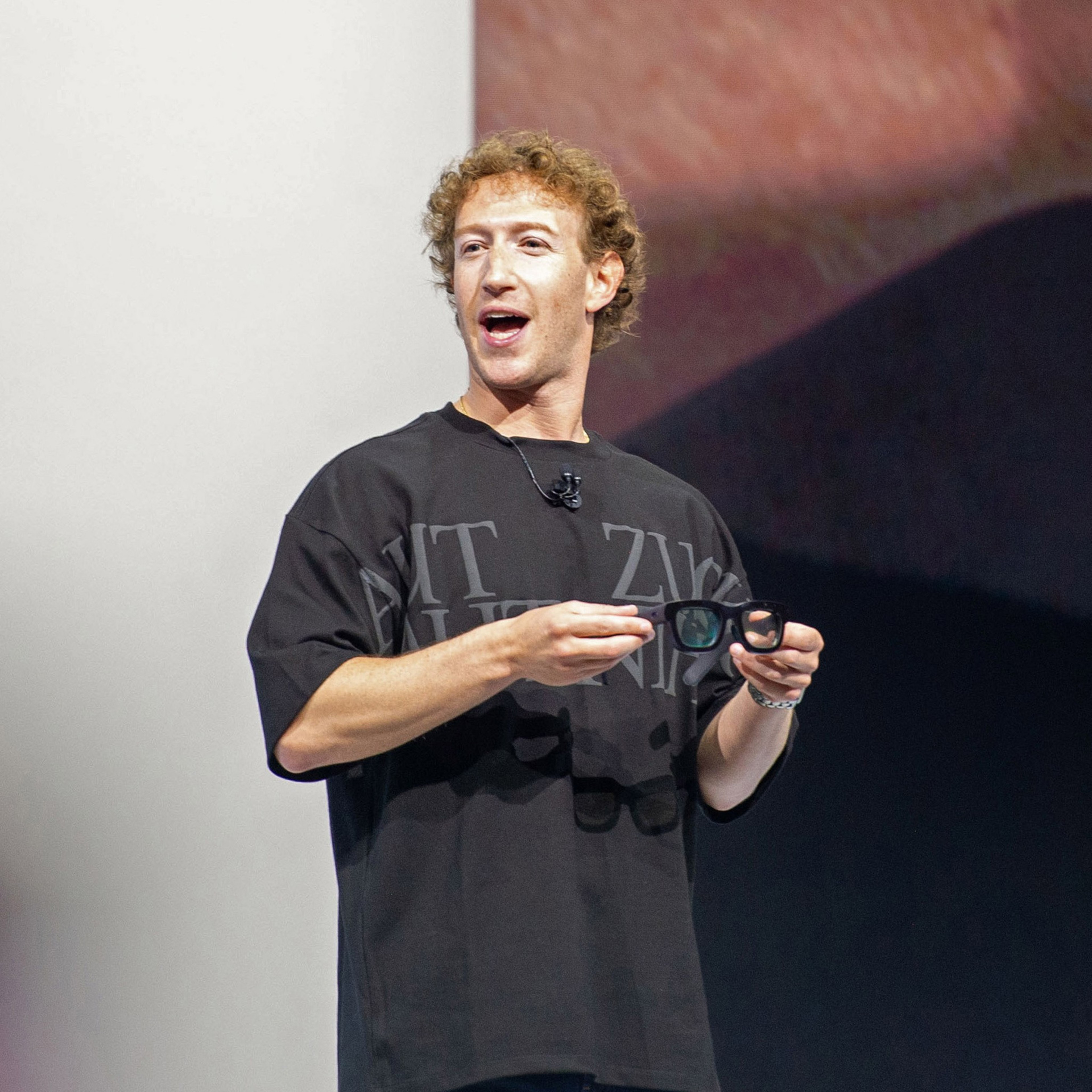 A man with curly hair wears a loose black shirt and holds a pair of dark-framed glasses while speaking on stage with a microphone clipped to his shirt.
