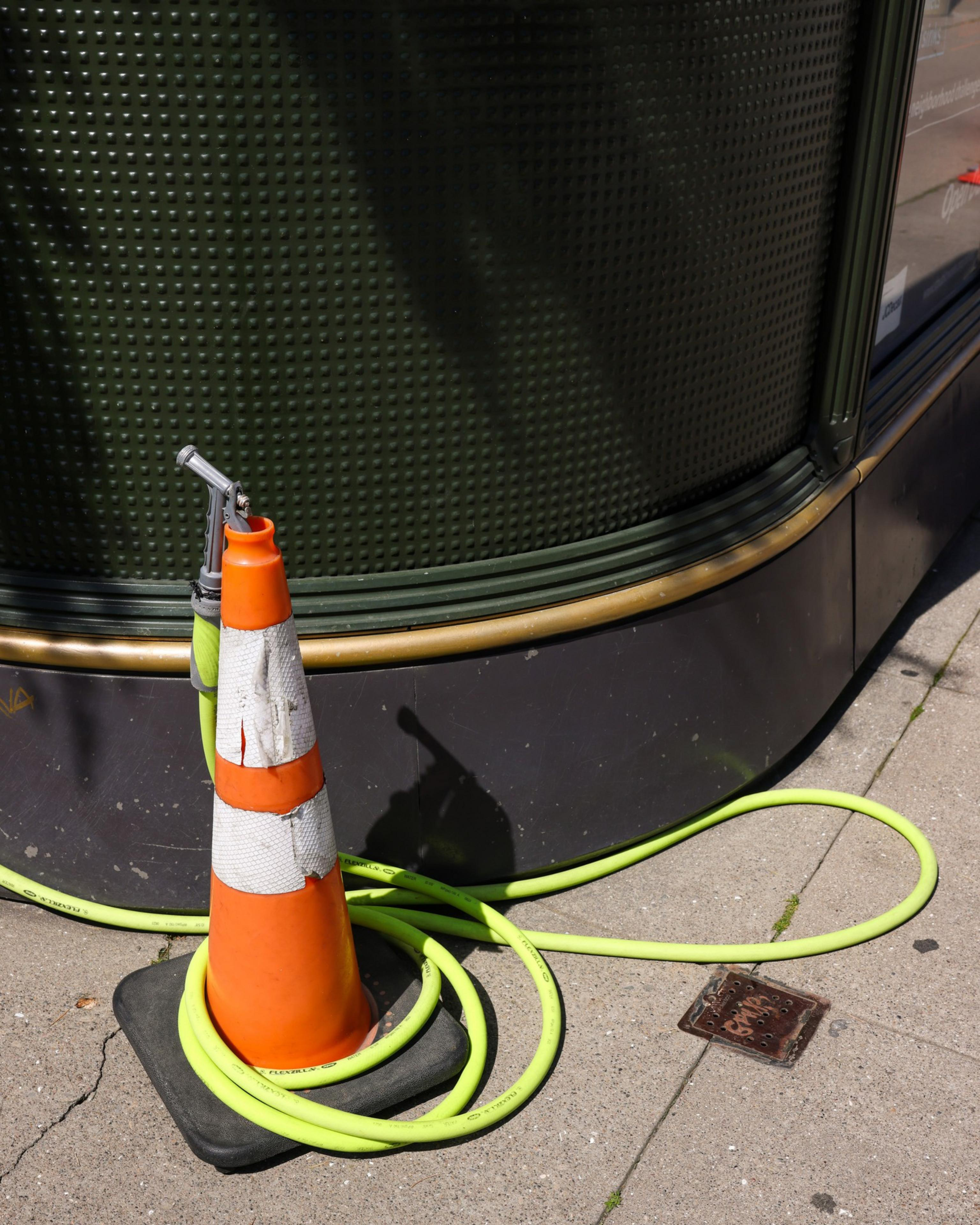 A bright orange traffic cone with white reflective stripes stands on a concrete sidewalk, with a lime green hose wrapped around its base, near a textured green wall.