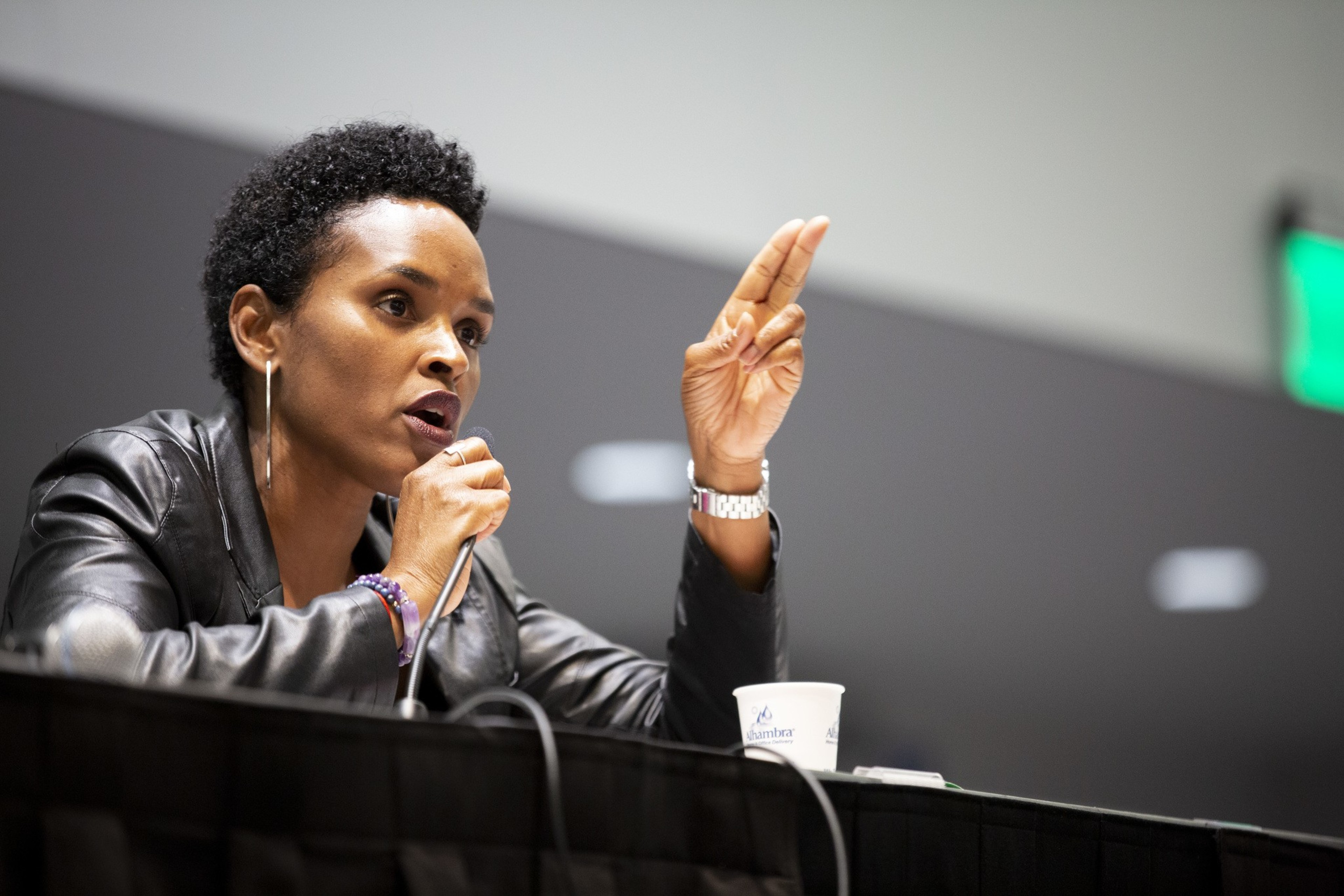 A person with short curly hair speaks into a microphone while gesturing with two fingers, wearing a leather jacket and hoop earrings, sitting at a panel table.