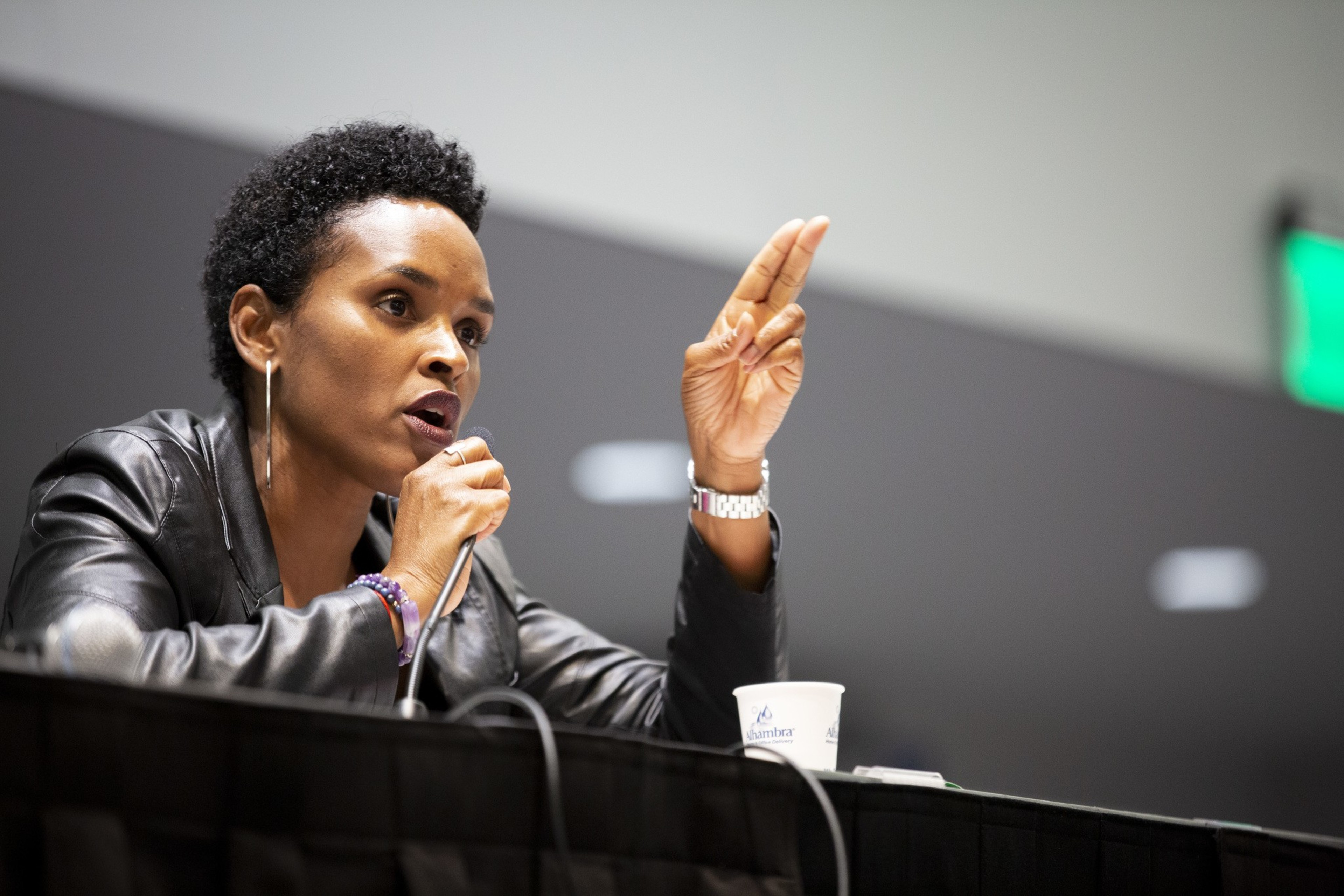 A person with short curly hair speaks into a microphone while gesturing with two fingers, wearing a leather jacket and hoop earrings, sitting at a panel table.
