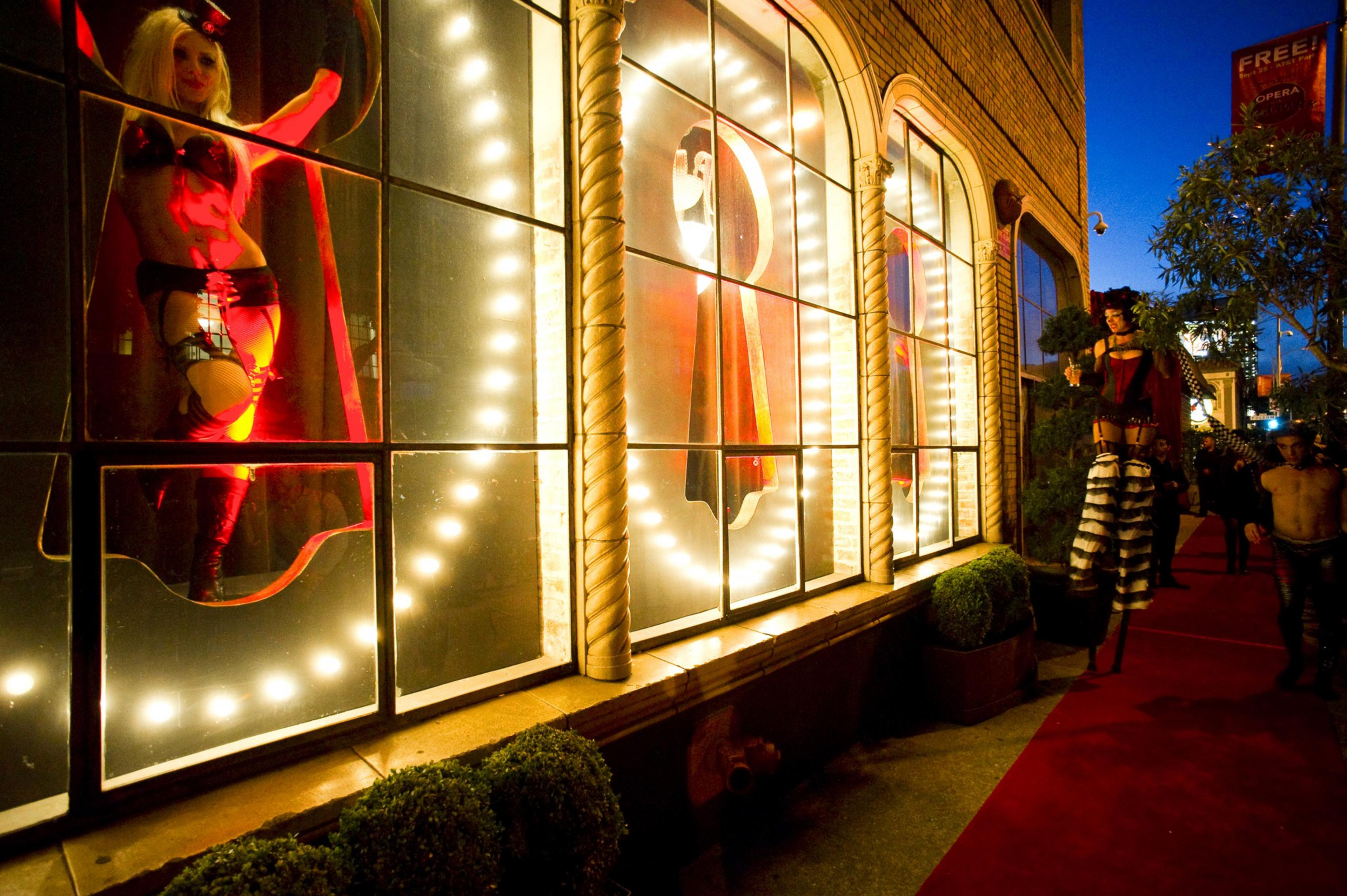 A woman in revealing attire poses in a brightly lit window, while another performer on stilts walks along a nighttime street with a red carpet, surrounded by people.