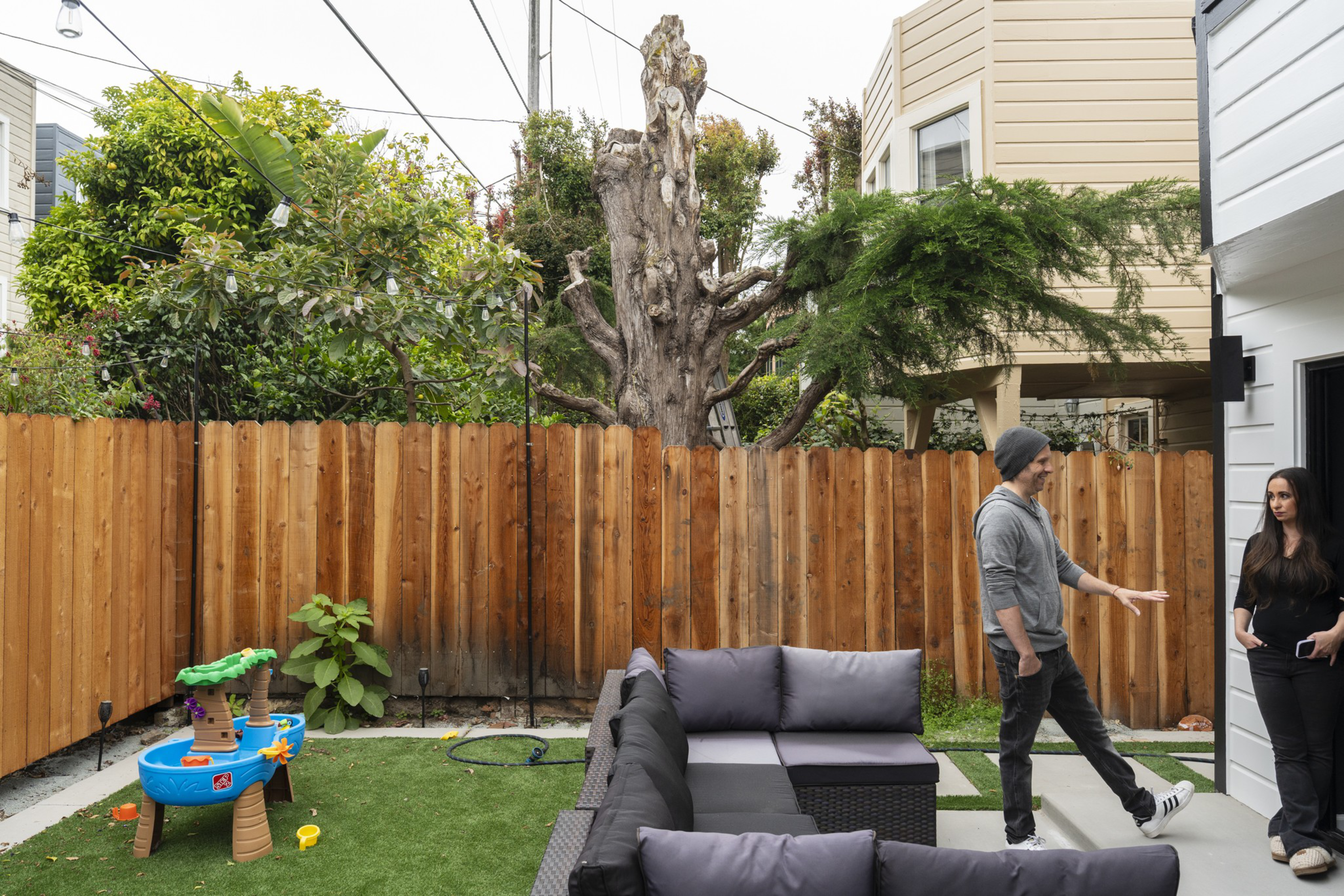 A backyard with artificial grass, a wooden fence, a water play table, outdoor seating, and two people talking near a house entrance.