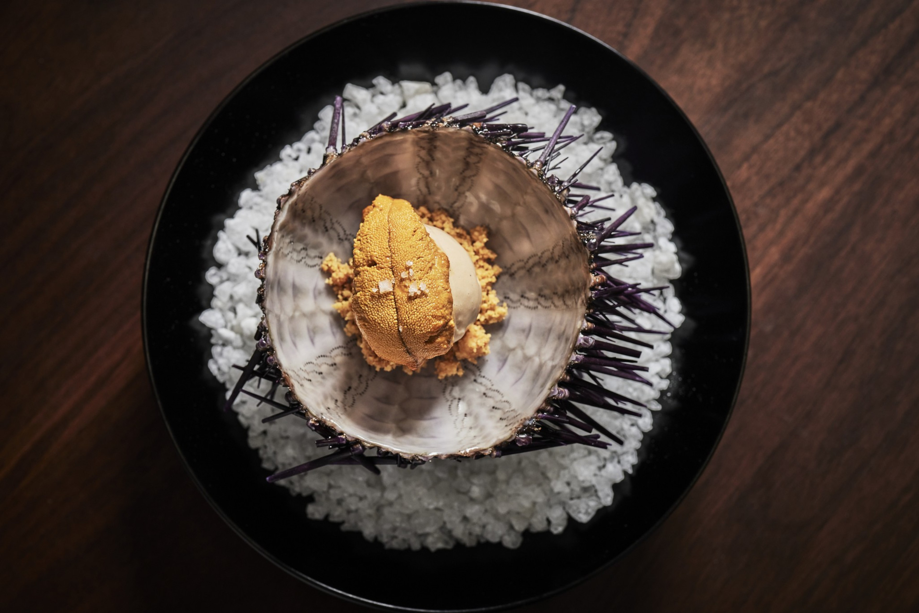 A spiky sea urchin shell is placed on coarse salt, containing golden sea urchin roe, in a black bowl on a wooden surface.