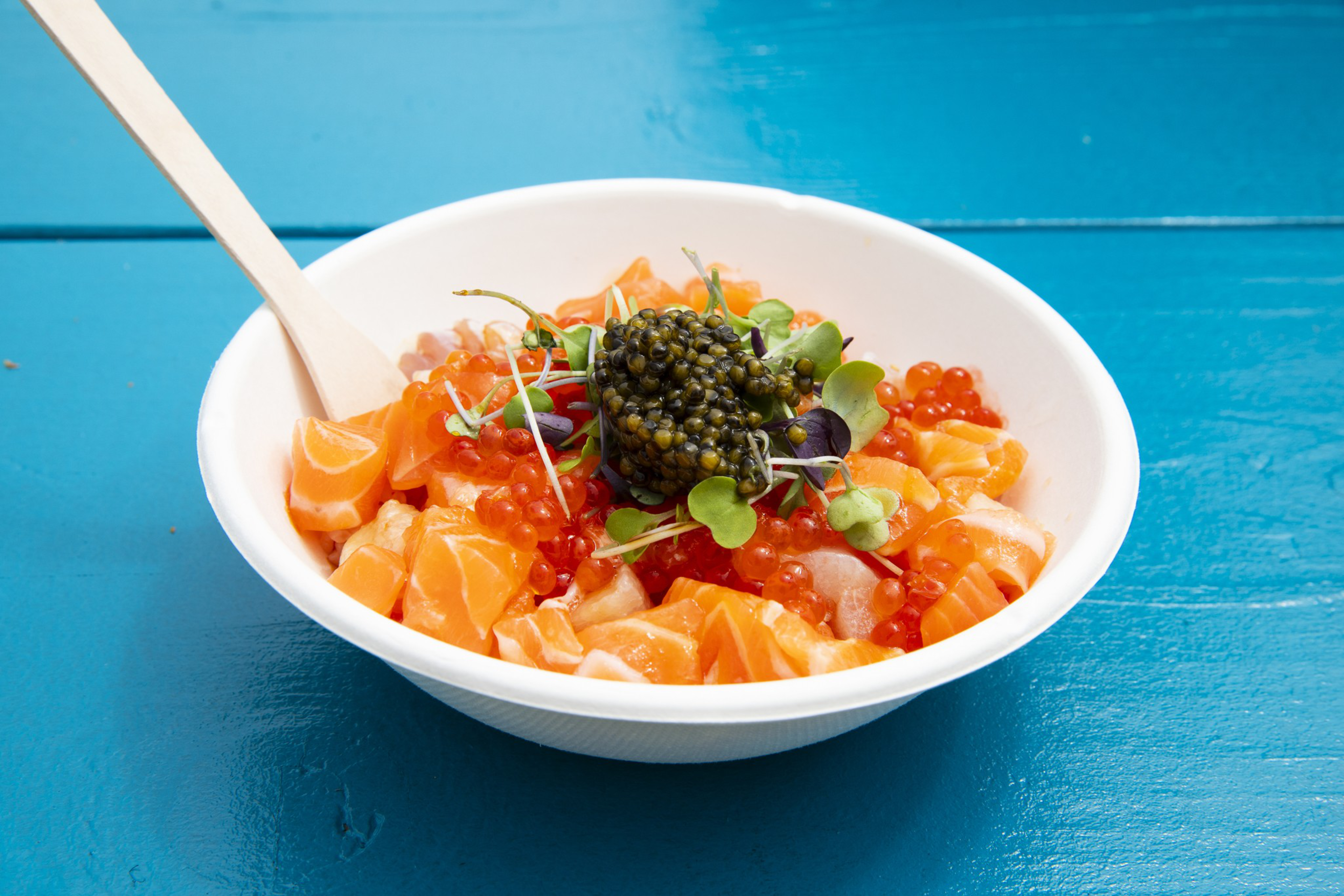 A white bowl contains diced salmon, red fish roe, green microgreens, and a dollop of black caviar, placed on a bright blue wooden surface with a wooden spoon.