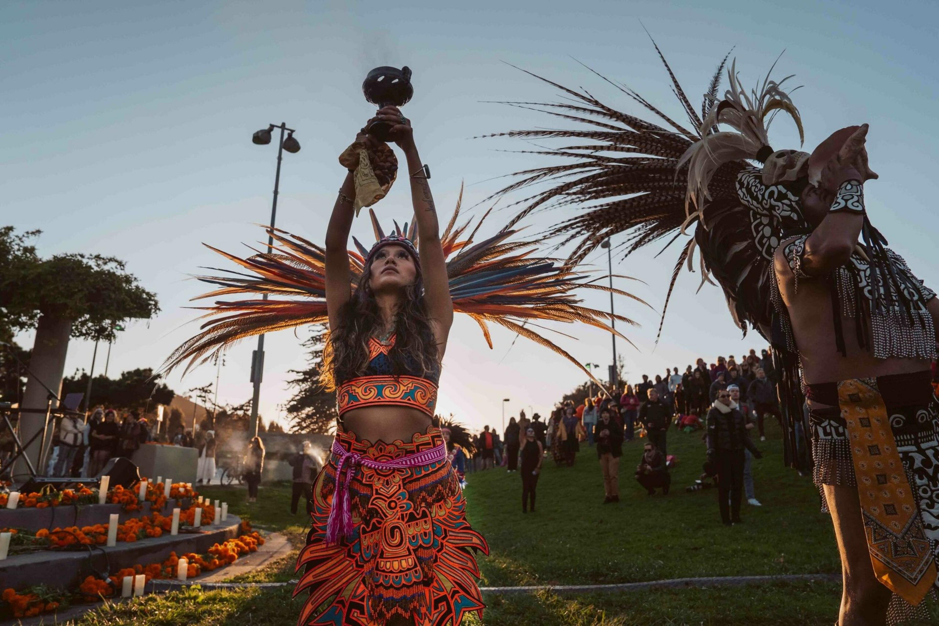 A woman in vibrant Aztec-style attire raises a vessel above her head, surrounded by people and an altar decorated with candles and marigolds at dusk.