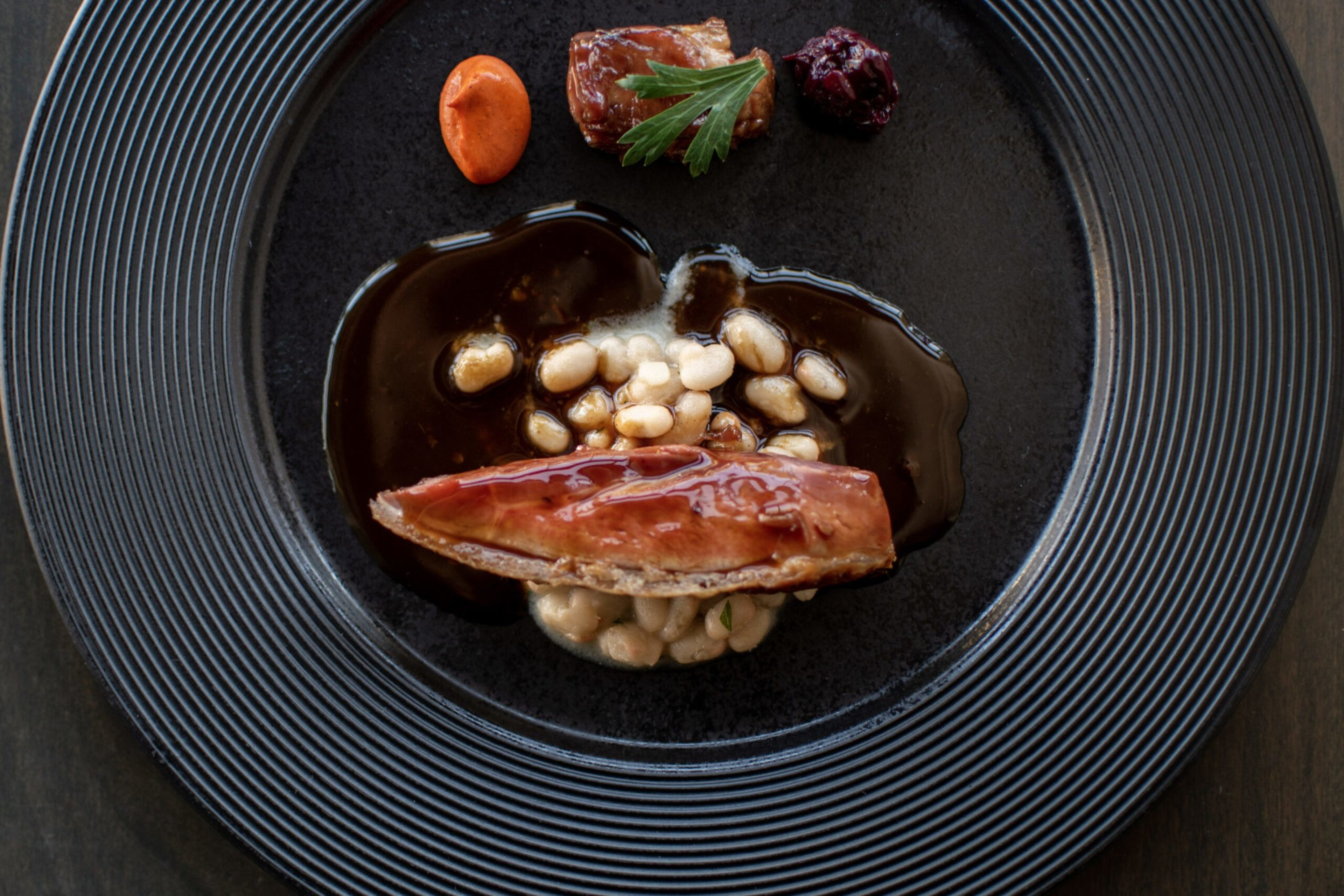 A black plate holds a piece of glazed fish atop white beans with dark sauce, alongside an orange dollop, a small meat cube with parsley, and a dark red garnish.